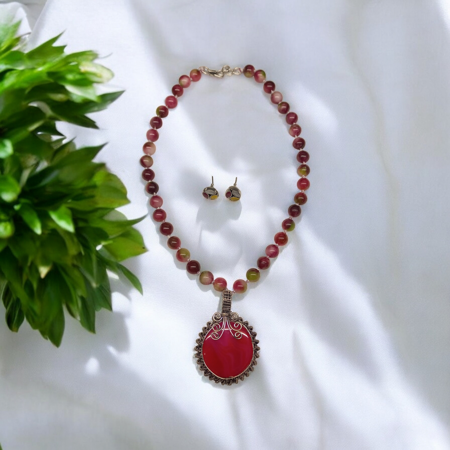 Beautiful Nature beads statement necklace with ear stud in dark pink colour placed on a white fabric with few green leaves to decorate the show. Best for casual and party wear.
