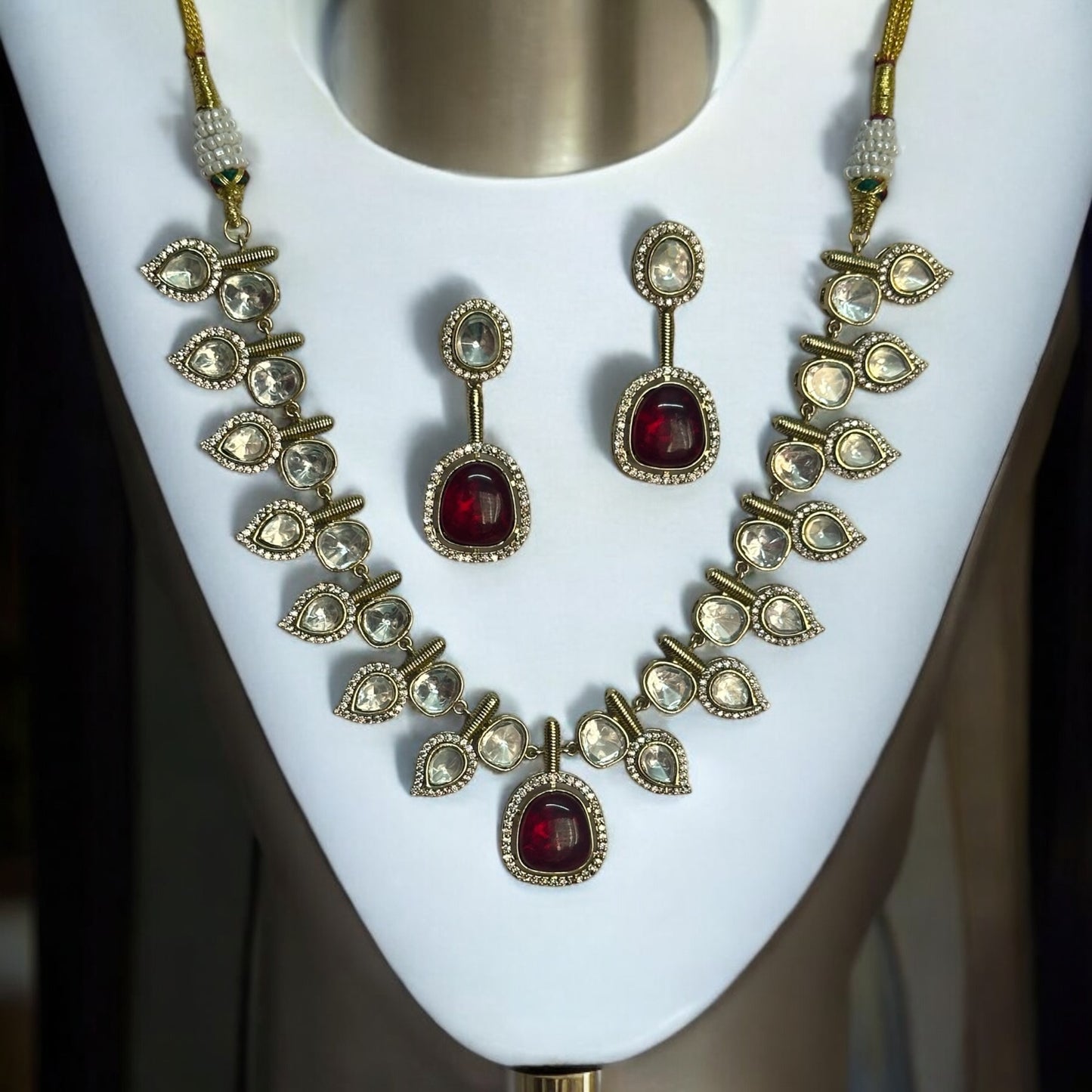 Elegant designer polki kundan necklace with teardrop accents and red gemstone centerpiece, paired with matching earrings. Jewellery set is placed on a white jewellery display stand.