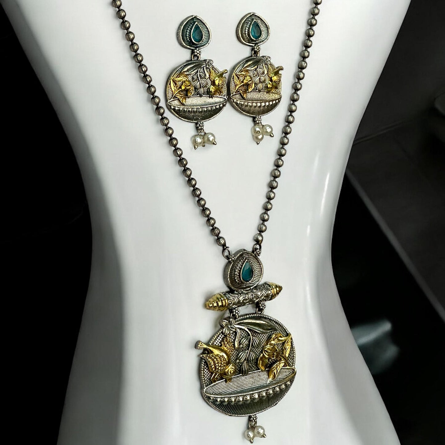 Dual tone German silver chain pendant set with gold floral and fish motifs, green stone accents, pearl drops, and matching earrings displayed on white jewellery stand