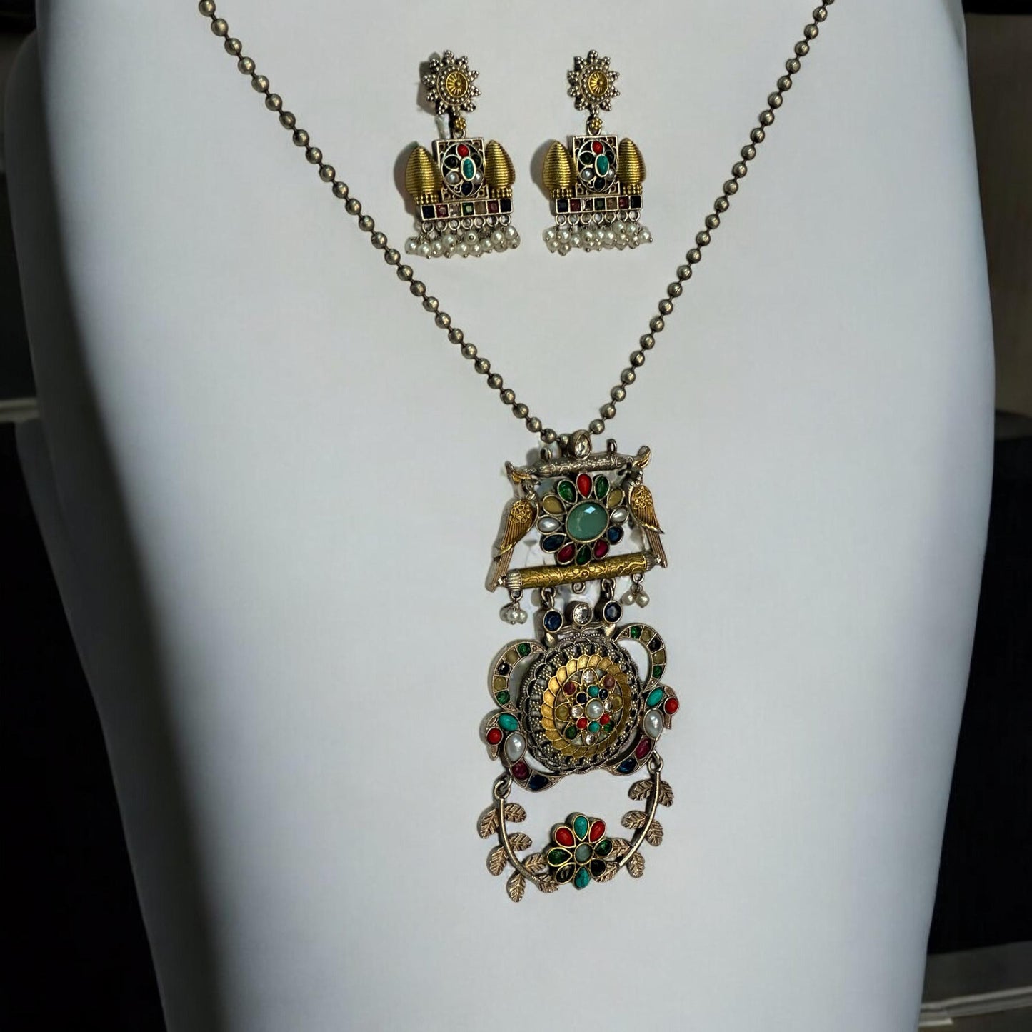 Dual Tone Apala Pachi Kundan Pendant Set with Navaratna Stones and Matching Earrings placed on a white coloured jewellery display stand.