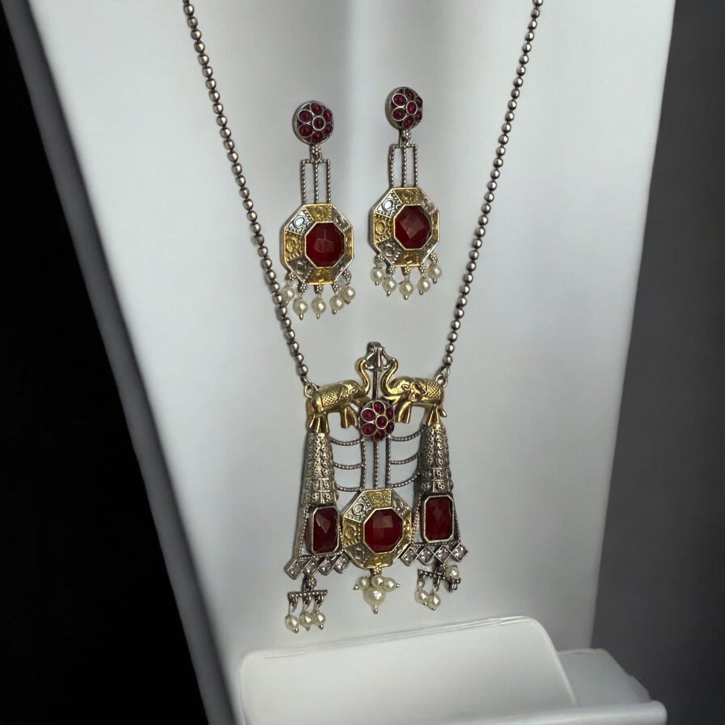 Side view of Dual Tone Silver Replica Necklace Set in ruby red and golden color featuring a detailed pendant with ruby red stones and hanging pearls, displayed on a white mannequin with a soft black and white gradient background.