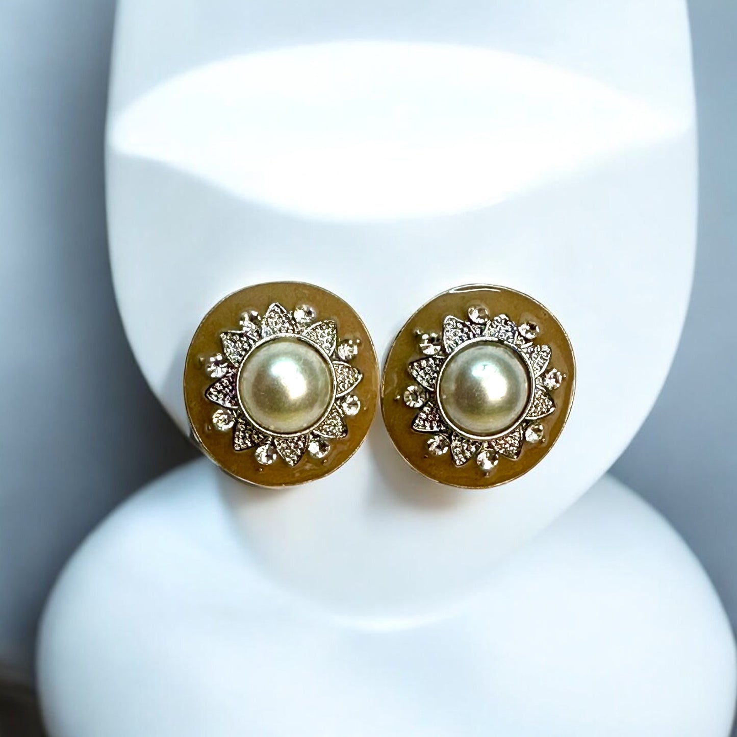 Elegant pearl stud earrings with a gold-toned circular base, featuring intricate floral detailing and embedded white stones, displayed on a white jewellery display stand with a soft blue backdrop.
