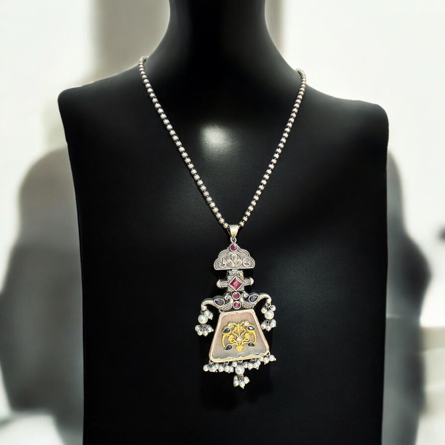German silver replica necklace with an intricate pendant design featuring floral motifs in gold and pink highlights, displayed on a black mannequin with a soft, elegant background.
