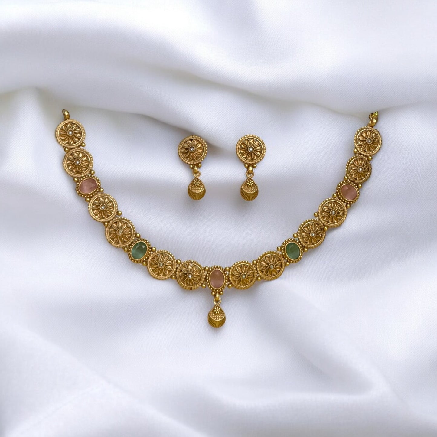 Gold-plated Rajwadi necklace set with intricate circular motifs and pastel green and pink accents, paired with matching gold-plated earrings, displayed on a white fabric..