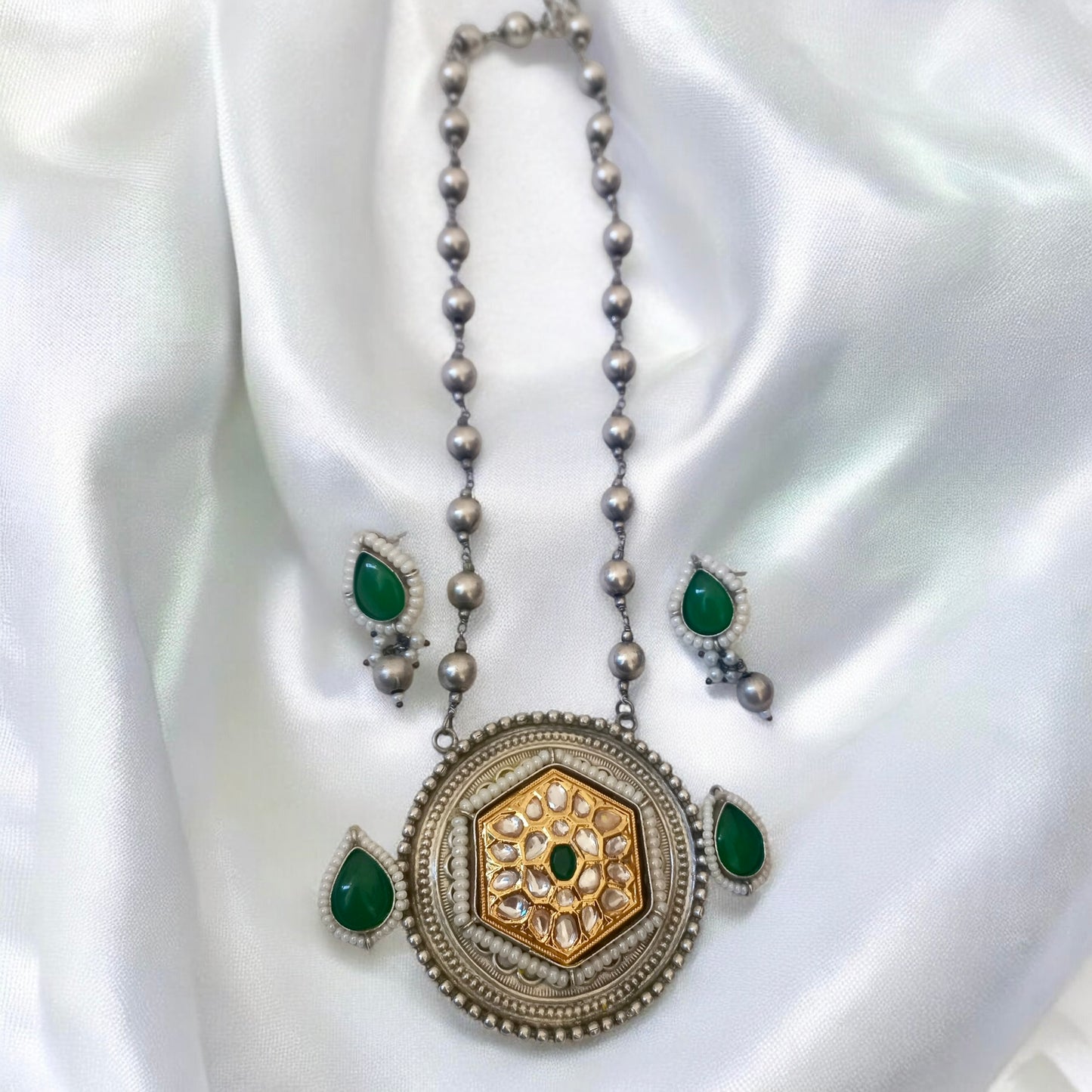 Long Dual Tone Apala Big Size Pendant Set in Green featuring an intricately designed silver-gold pendant with Kundan work, green stone accents, and matching teardrop earrings. Perfect for ethnic and festive wear. Displayed on a white colored fabric.