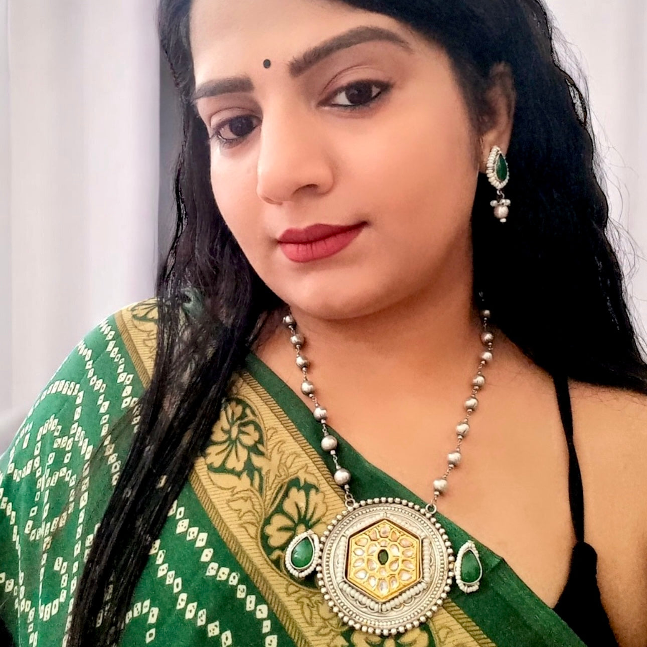A model wearing and displaying a Long Dual Tone Apala Big Size Pendant Set in Green featuring an intricately designed silver-gold pendant with Kundan work, green stone accents, and matching teardrop earrings. Perfect for ethnic and festive wear.