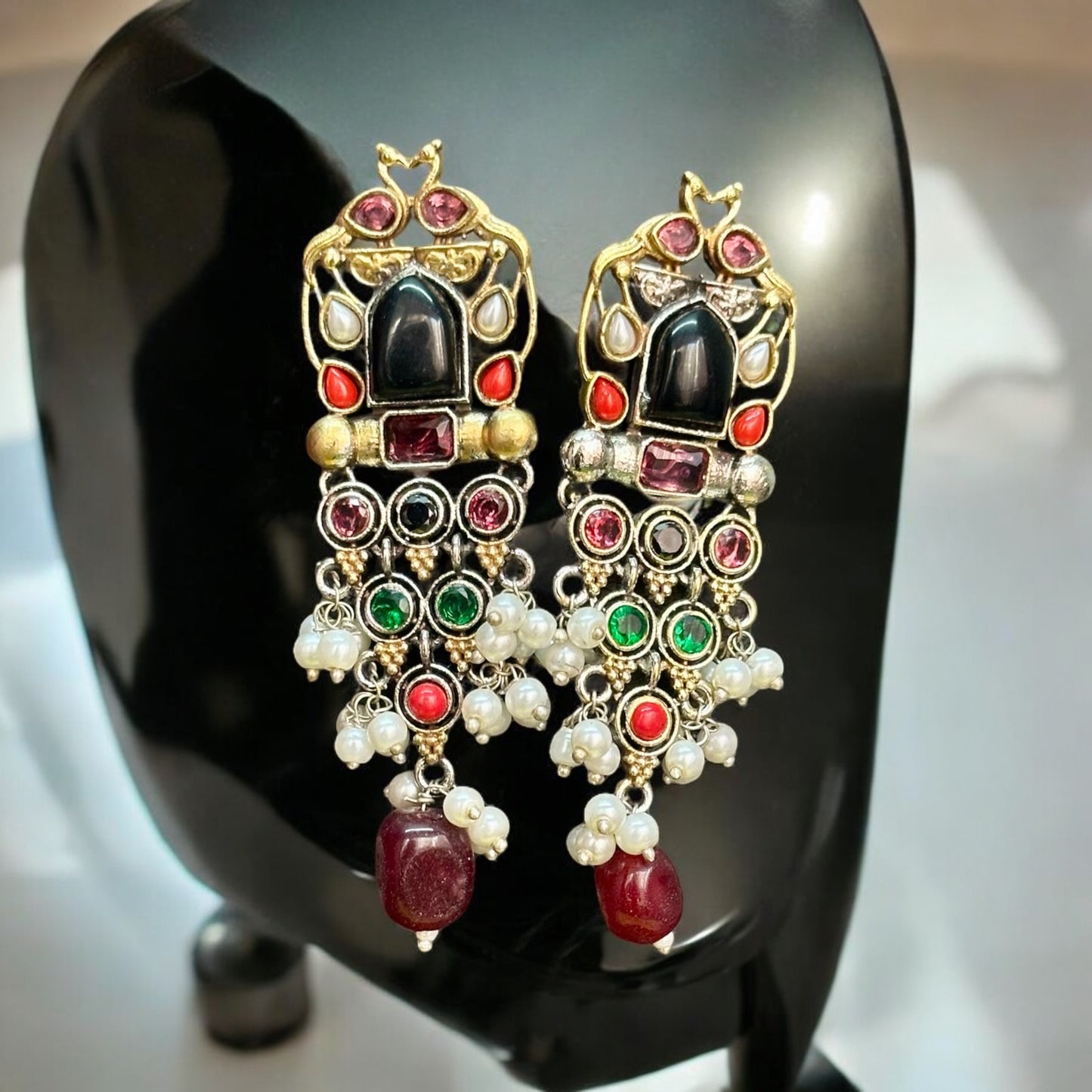 Long silver dual-tone earrings with intricate detailing, adorned with multicolor stones, pearls displayed against a soft black jewellery display stand.