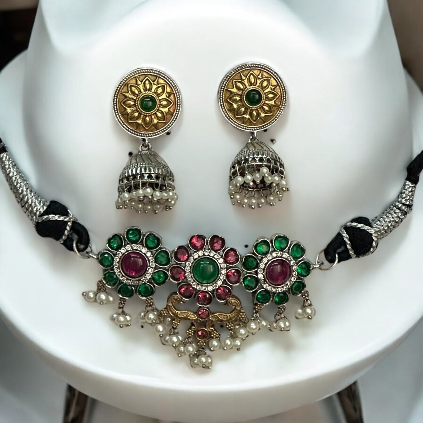 Minimal silver dual-tone choker featuring intricate floral motifs with green and pink gemstones, accented with pearls. Paired with gold-toned round jhumka earrings with detailed patterns, displayed on a white background.
