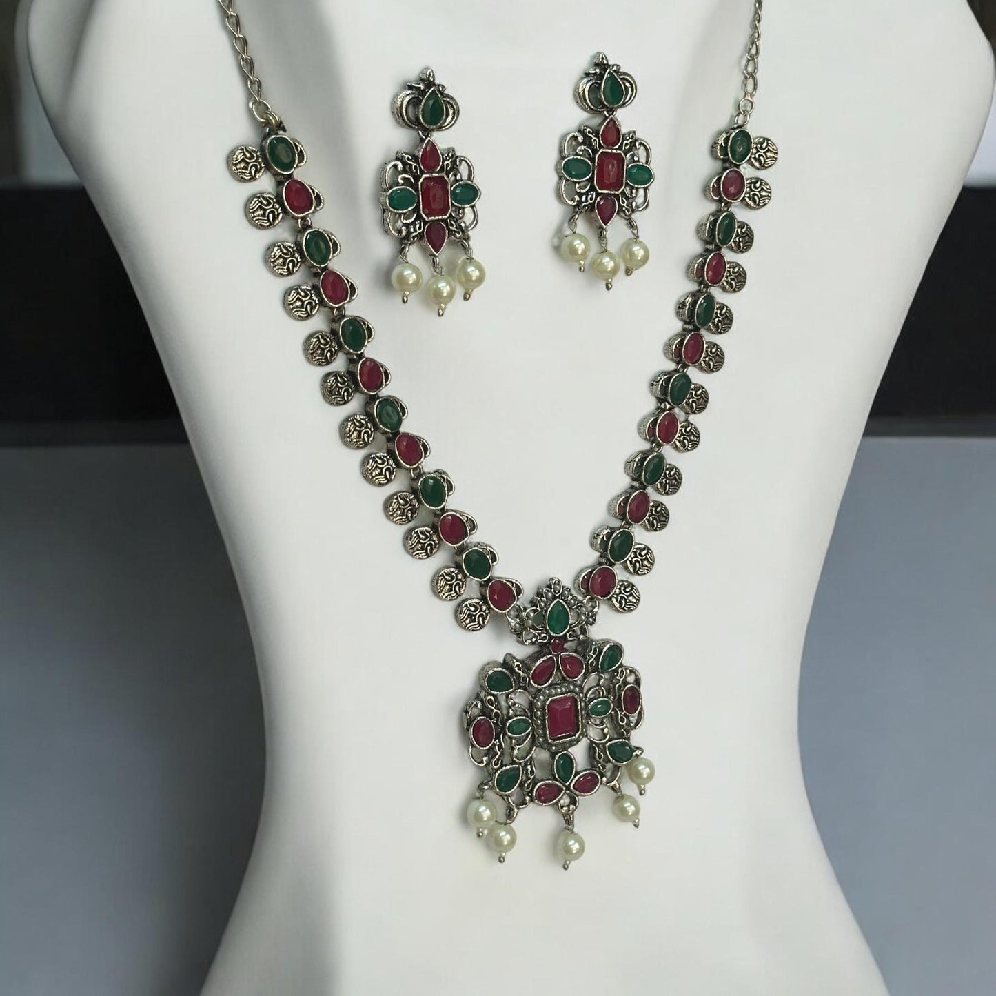 Side view of Oxidised necklace set featuring ruby-red and emerald-green stones with pearl drops, paired with matching earrings, displayed on a white mannequin against a soft background.