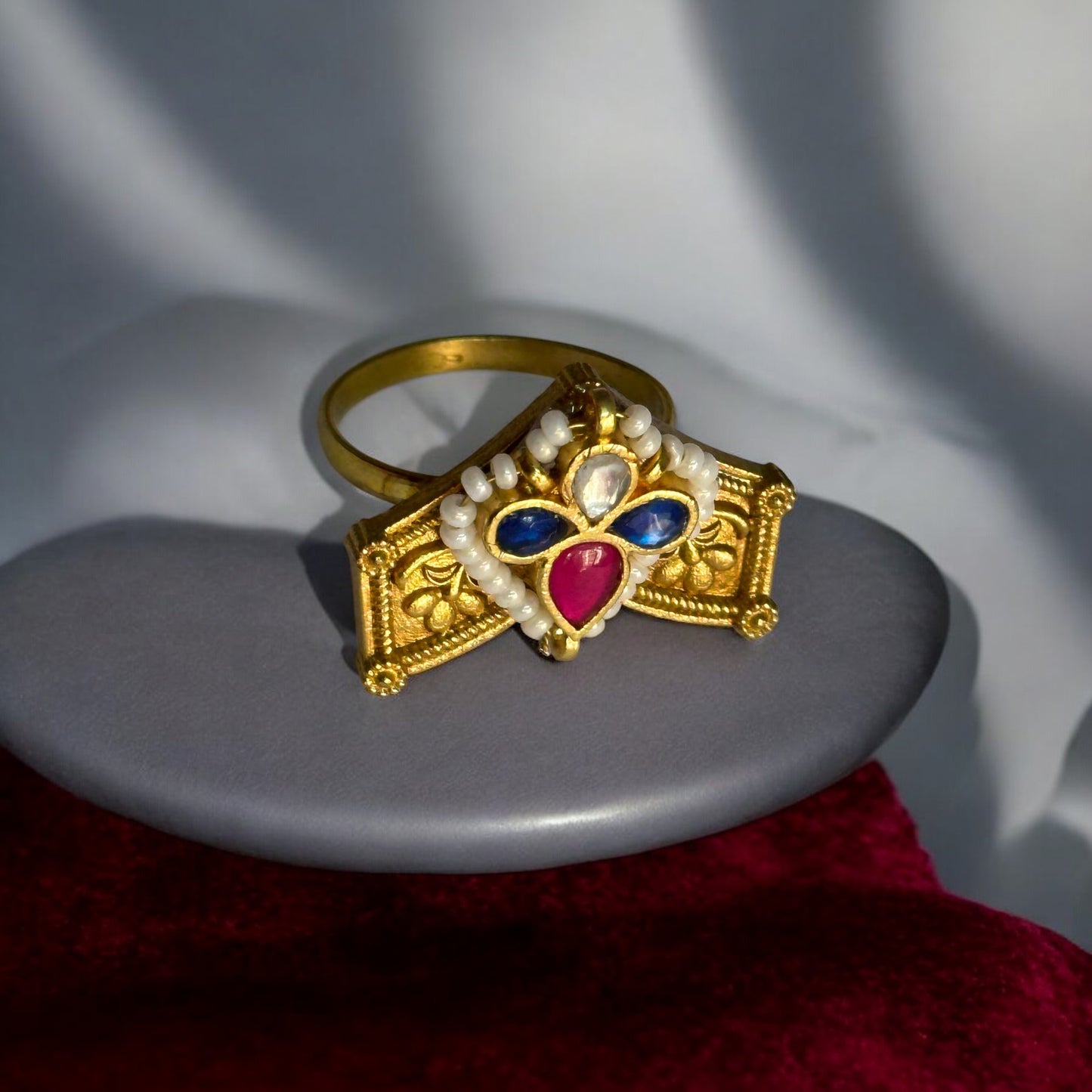 Exquisite Pachi Kundan finger ring in gold-tone with intricate detailing, featuring red, blue, and white gemstones and delicate pearl accents. Placed on a grey display stand.