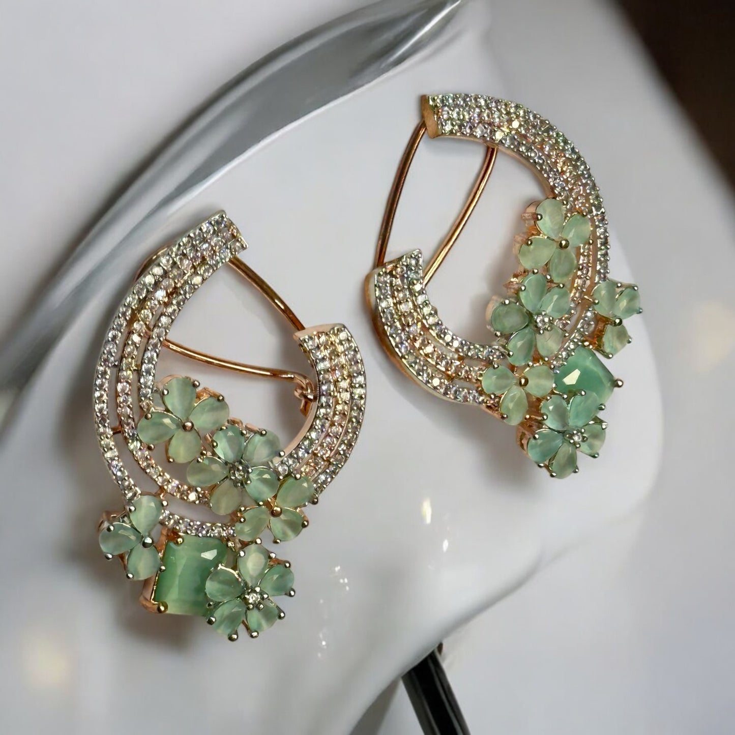 Partywear Statement American Diamond Earrings – crescent-shaped design with mint floral accents, gemstones, and layers of American diamonds. It is placed on a white jewellery display stand. Ideal for festive and formal occasions.