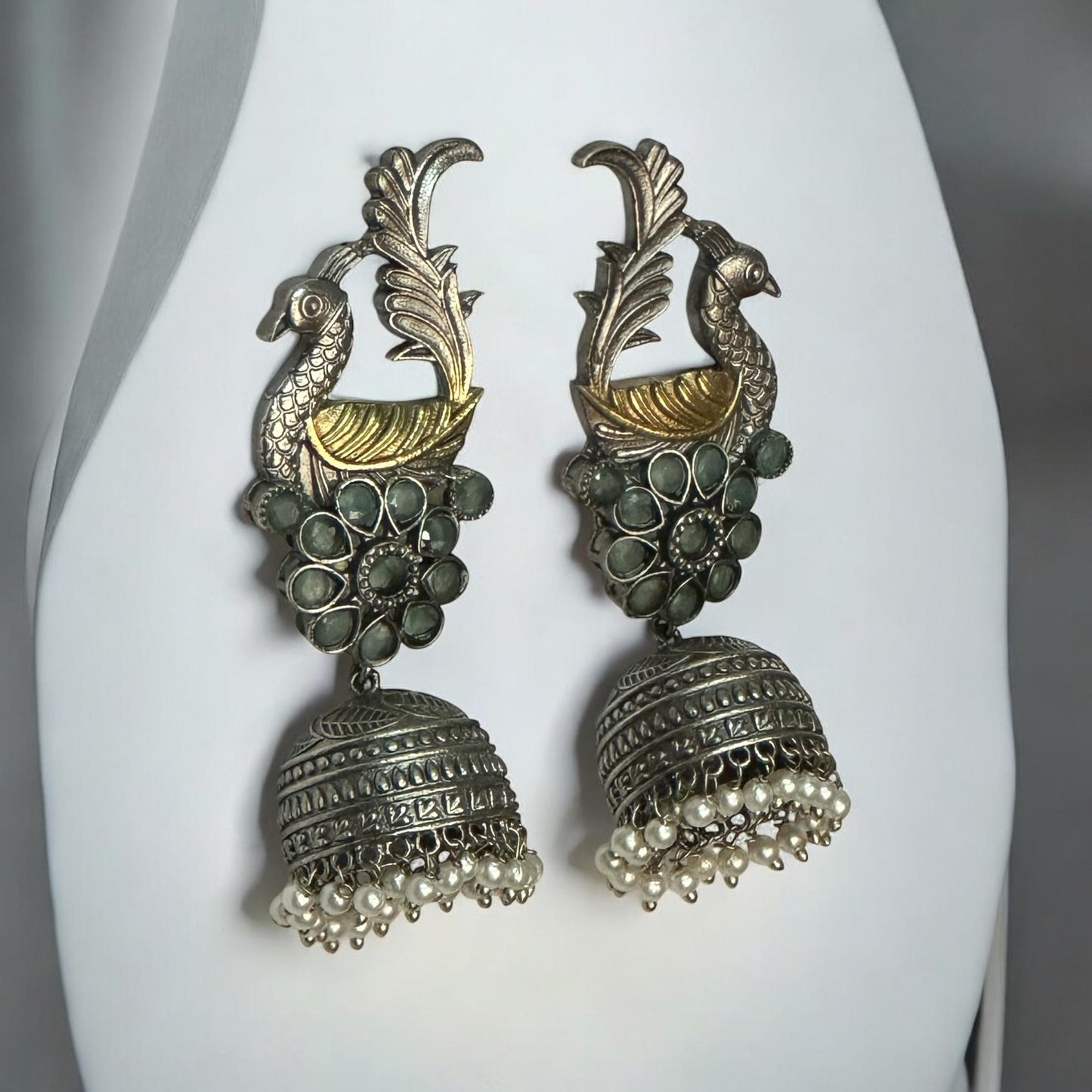 Dual-tone peacock jhumka earrings with floral stone detailing and pearl drops displayed on white jewellery stand – Bella & Diva