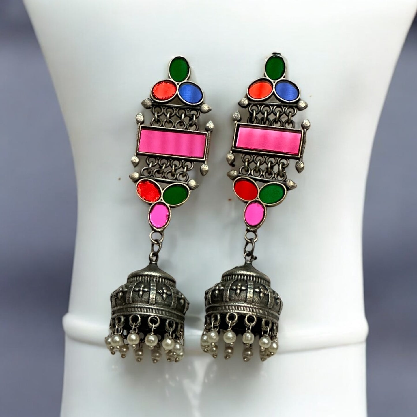 German silver earrings with colorful glass inlays in pink, green, blue, and red, featuring a jhumka design with pearl accents placed on a white jewellery display stand.
