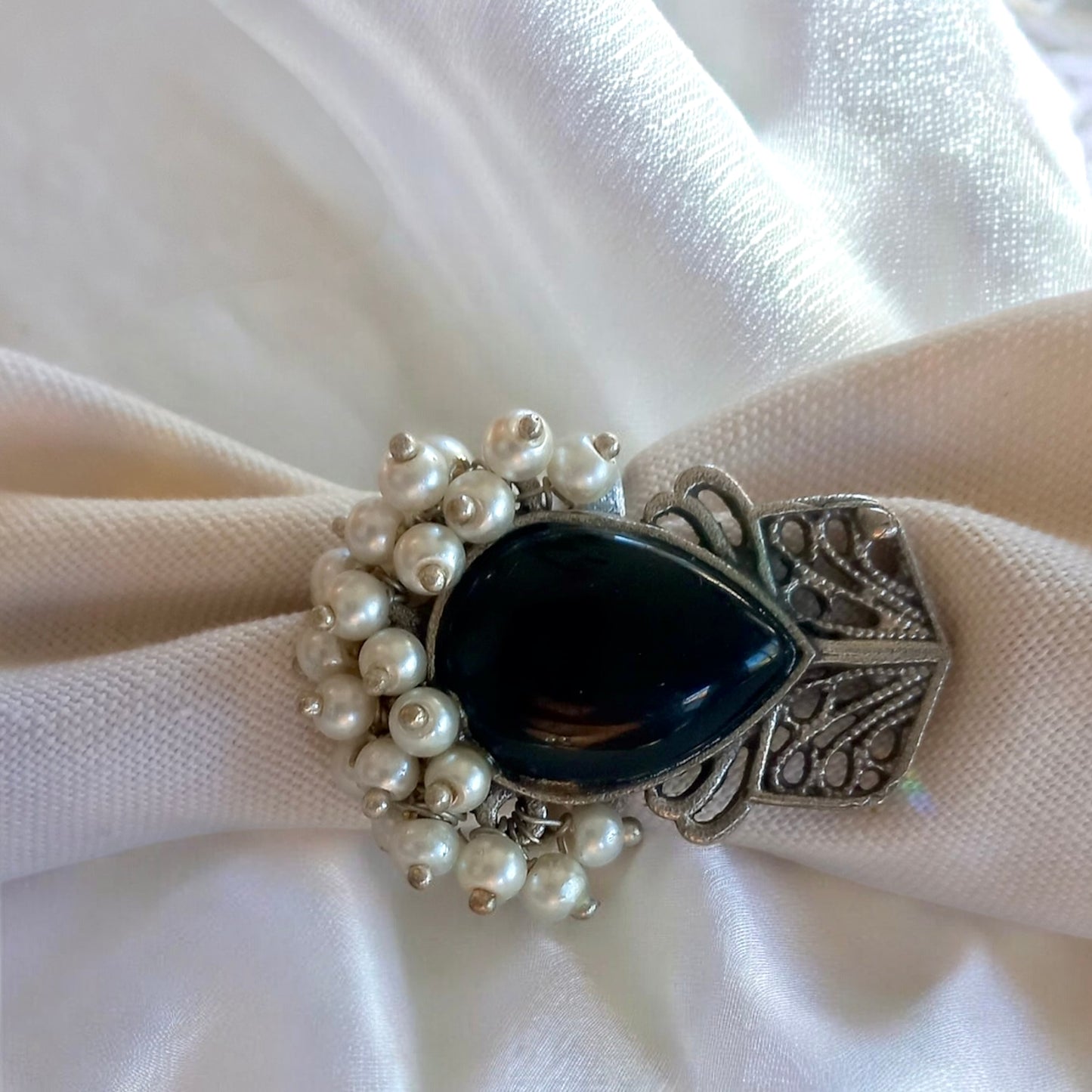 A white fabric backgroundi wth a Silver Replica Black Ring, showcasing a large teardrop-shaped black stone surrounded by white pearl-like beads and vintage silver detailing, set against a satin background.