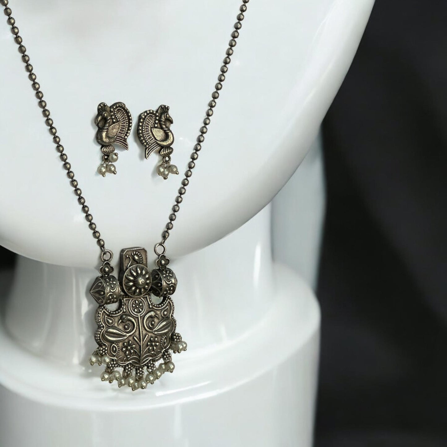 Close look of Silver replica designer chain pendant set in antique peacock design with pearl drops and matching earrings displayed on white jewellery stand