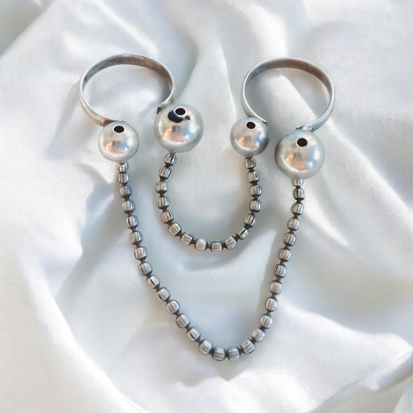 A fabric background with a Silver Replica Adjustable Double Finger Ring, featuring silver-toned chain links and pearl-like accents draped across two fingers, creating a bold fashion statement.