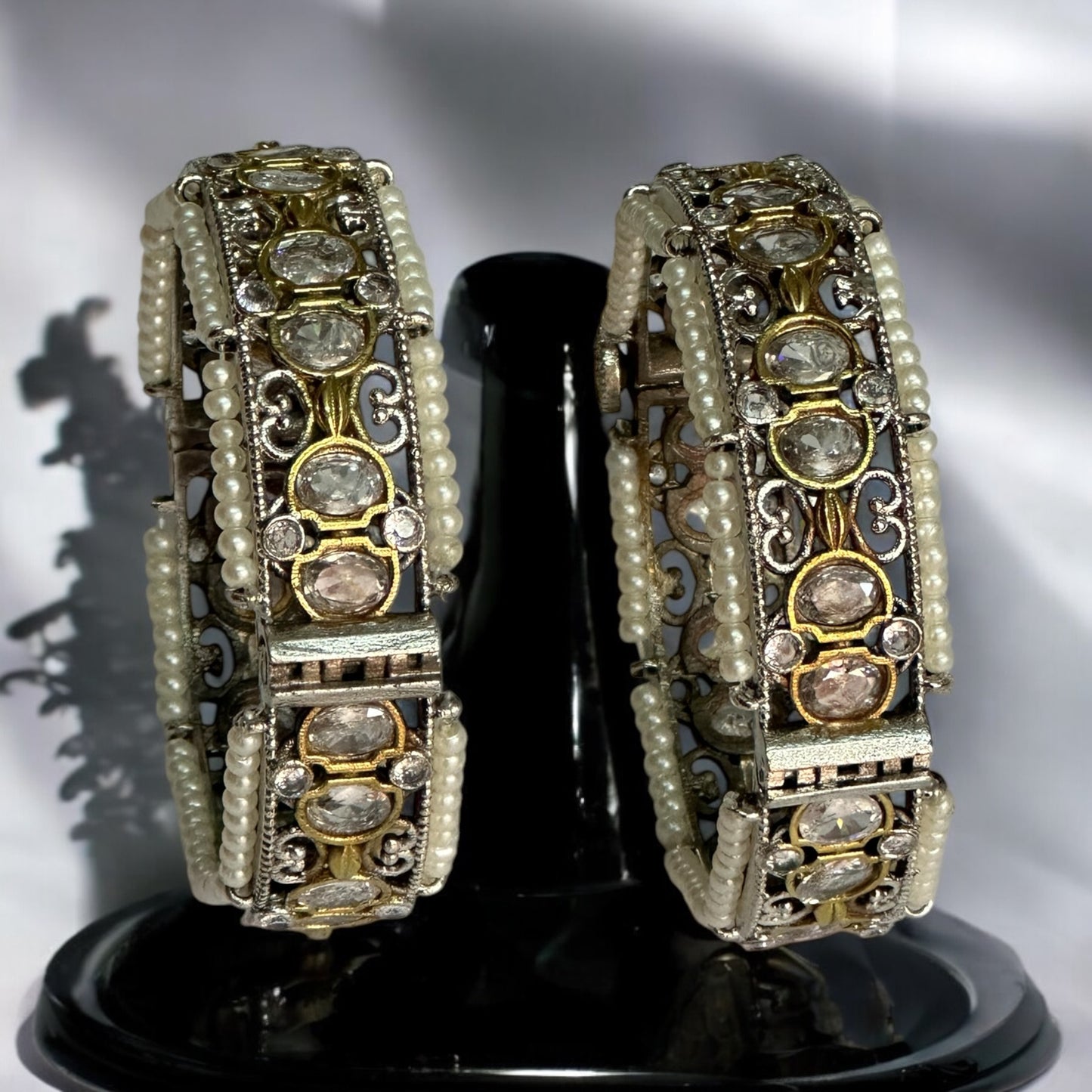 Elegant silver replica bangles adorned with intricate white pearl detailing and sparkling stonework. Its placed on a black coloured jewellery display stand.