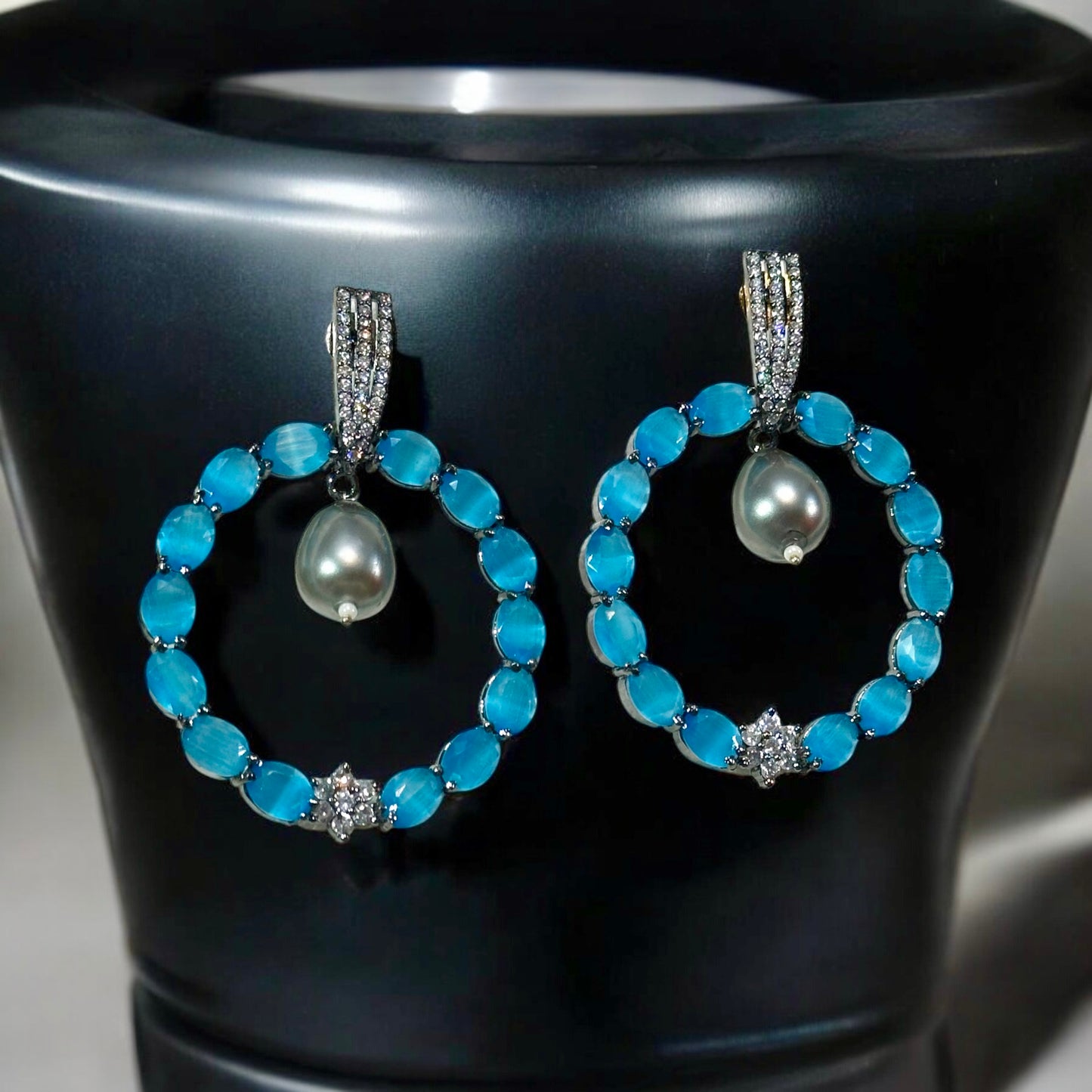 Statement American diamond earrings featuring a circular blue stone design with a hanging pearl drop, perfect for partywear. Placed on a black jewellery display stand.