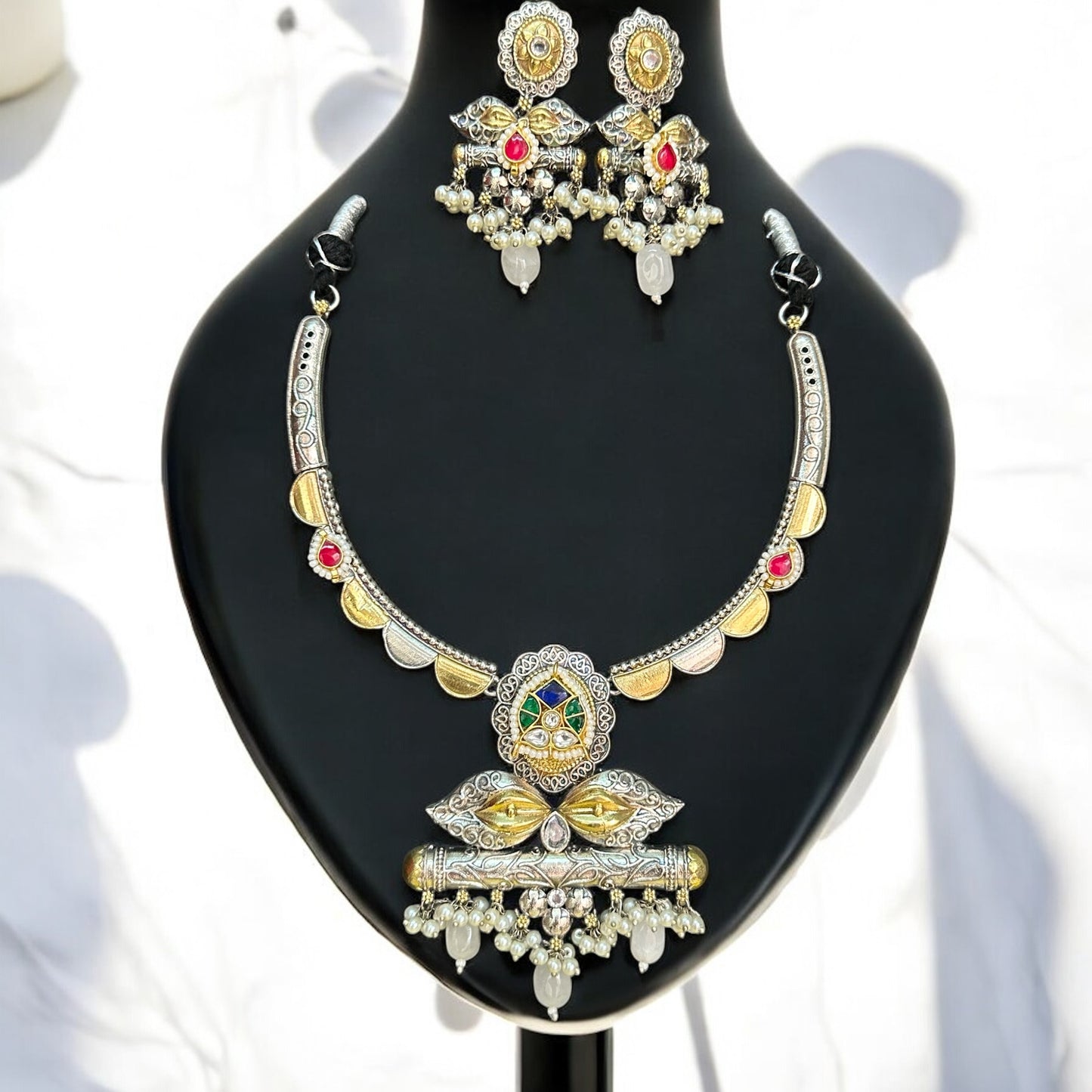 Amrapali dual-tone hasli necklace set featuring intricate pachi kundan and jadau work, adorned with vibrant pink, green, and white stones, complemented by matching statement earrings, displayed on a black mannequin with a soft sunlight backdrop.