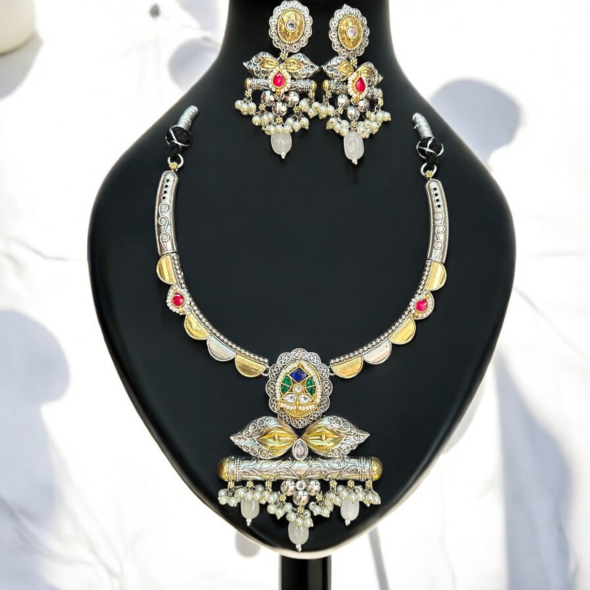Amrapali dual-tone hasli necklace set featuring intricate pachi kundan and jadau work, adorned with vibrant pink, green, and white stones, complemented by matching statement earrings, displayed on a black mannequin with a soft sunlight backdrop.