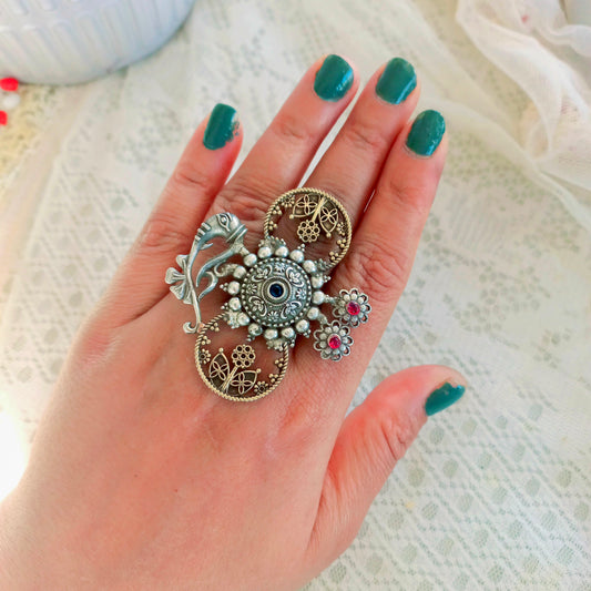 An Adjustable Fusion Oxidised Finger Ring featuring intricate oxidised silver detailing, floral filigree patterns, and delicate gemstone embellishments, worn on a hand with green-painted nails over a lace fabric background.