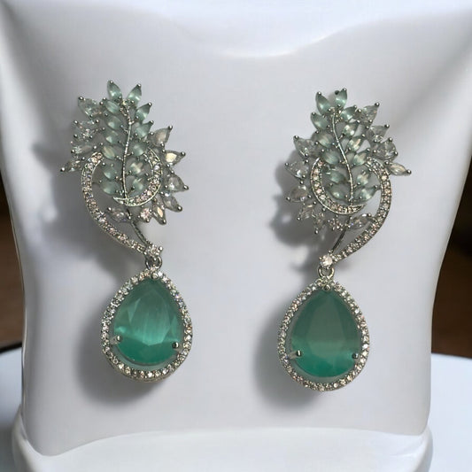 Beautiful American Diamond Statement Earrings In Greenish Magenta Shade placed on a white coloured jewellery display stand with a leaves design.