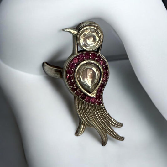 The image shows a bird-shaped ring. The ring is silver-colored with a gemstone forming the bird's head and a larger, teardrop-shaped gemstone surrounded by smaller red stones forming the bird's body. The bird's tail is stylized with several curved lines. 