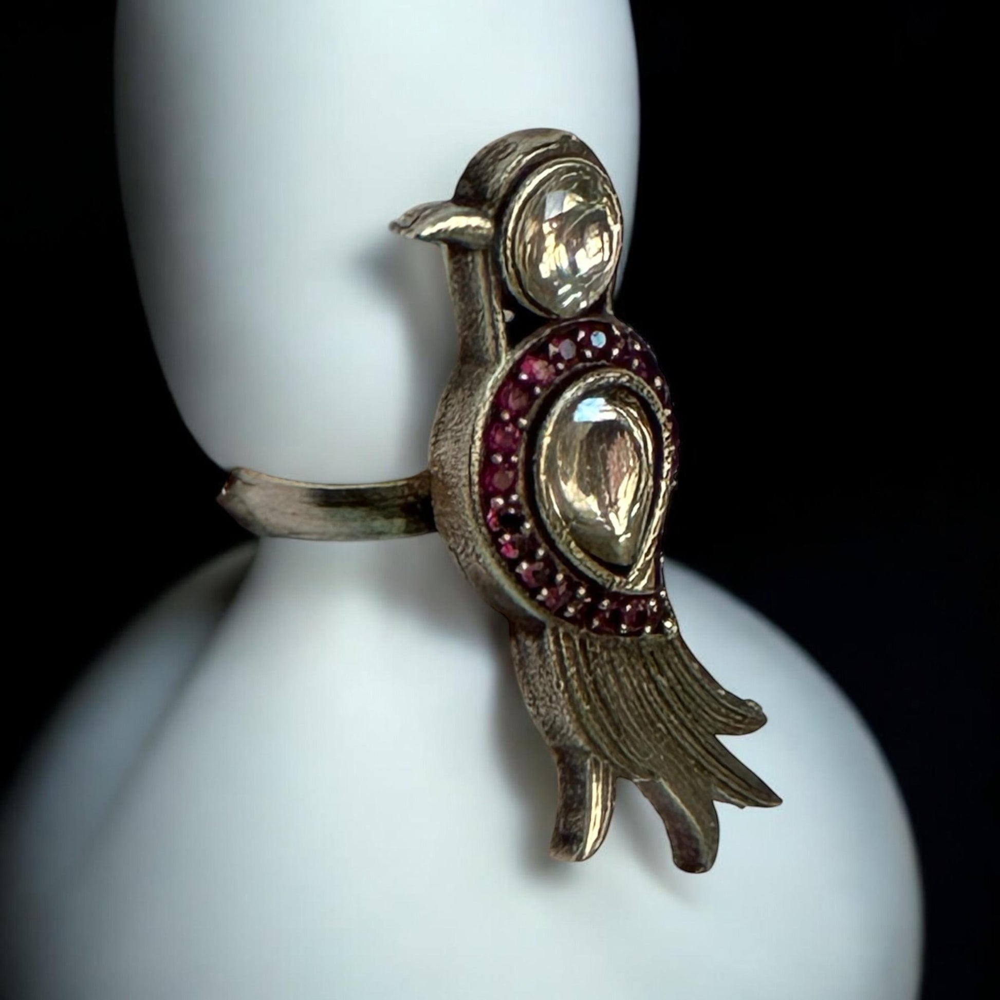 The image shows a bird-shaped ring. The ring is silver-colored with a gemstone forming the bird's head and a larger, teardrop-shaped gemstone surrounded by smaller red stones forming the bird's body. The bird's tail is stylized with several curved lines with white coloured background.