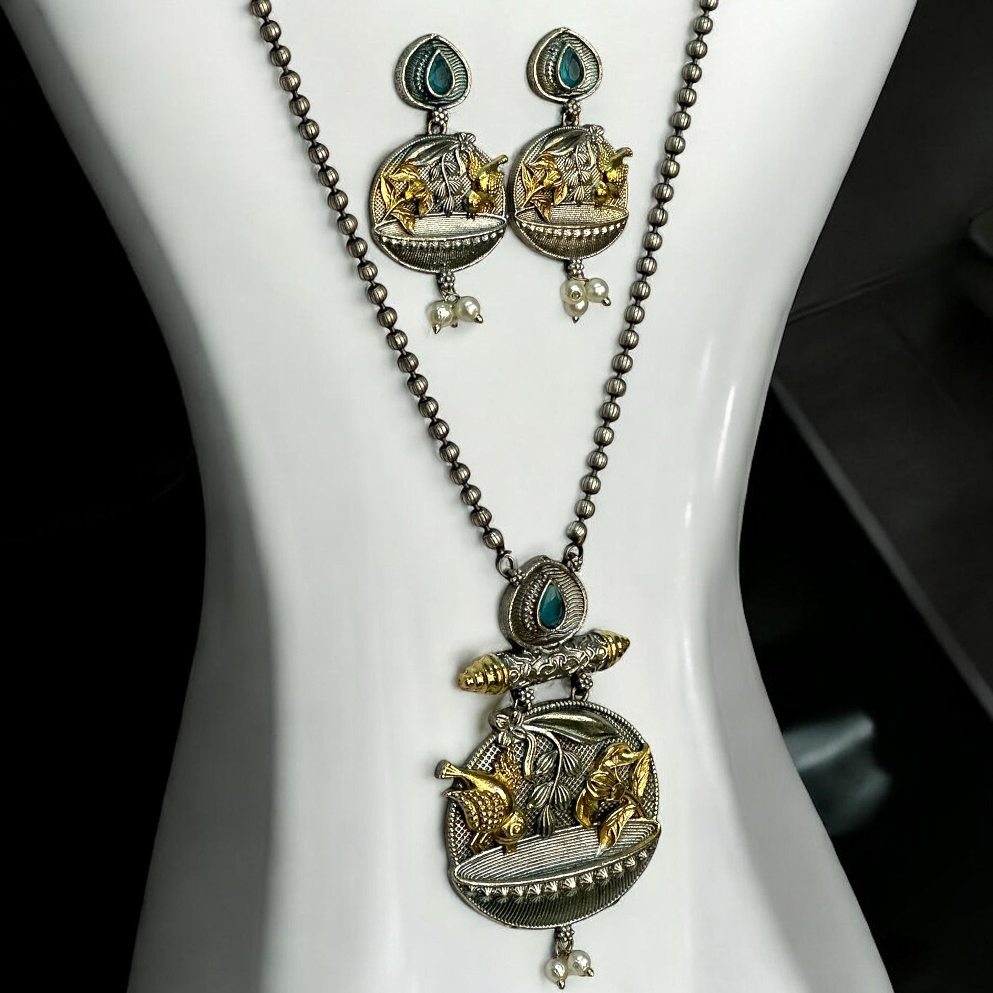 Dual tone German silver chain pendant set with gold floral and fish motifs, green stone accents, pearl drops, and matching earrings displayed on white jewellery stand