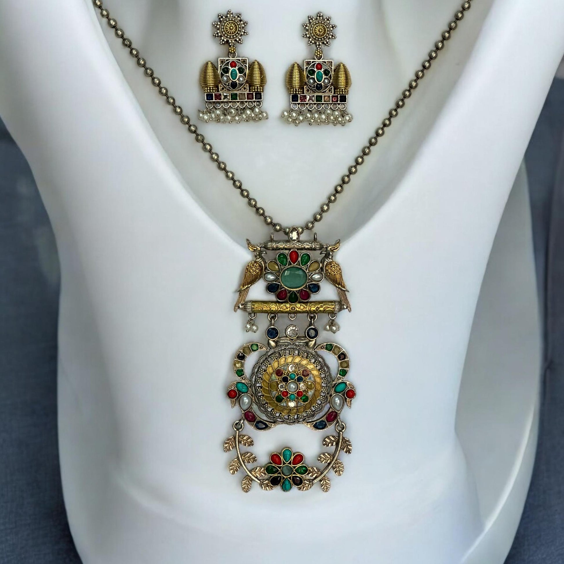 Dual Tone Apala Pachi Kundan Pendant Set with Navaratna Stones and Matching Earrings placed on a white coloured jewellery display stand.