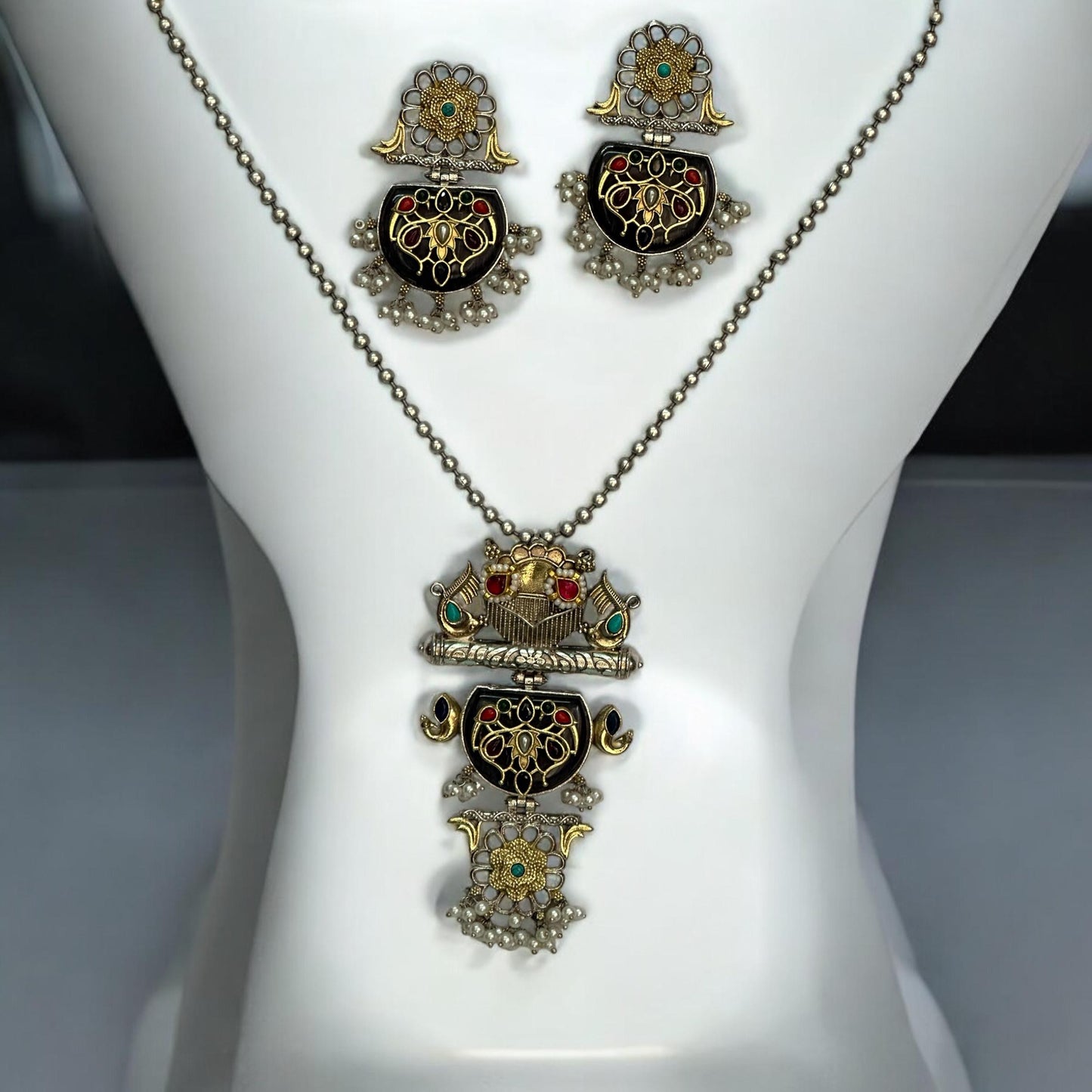 Dual Tone Apala Silver Long Pendant Set with Intricate Carving and Black Stone Finish, Featuring Matching Earrings placed on a white coloured jewellery display stand.