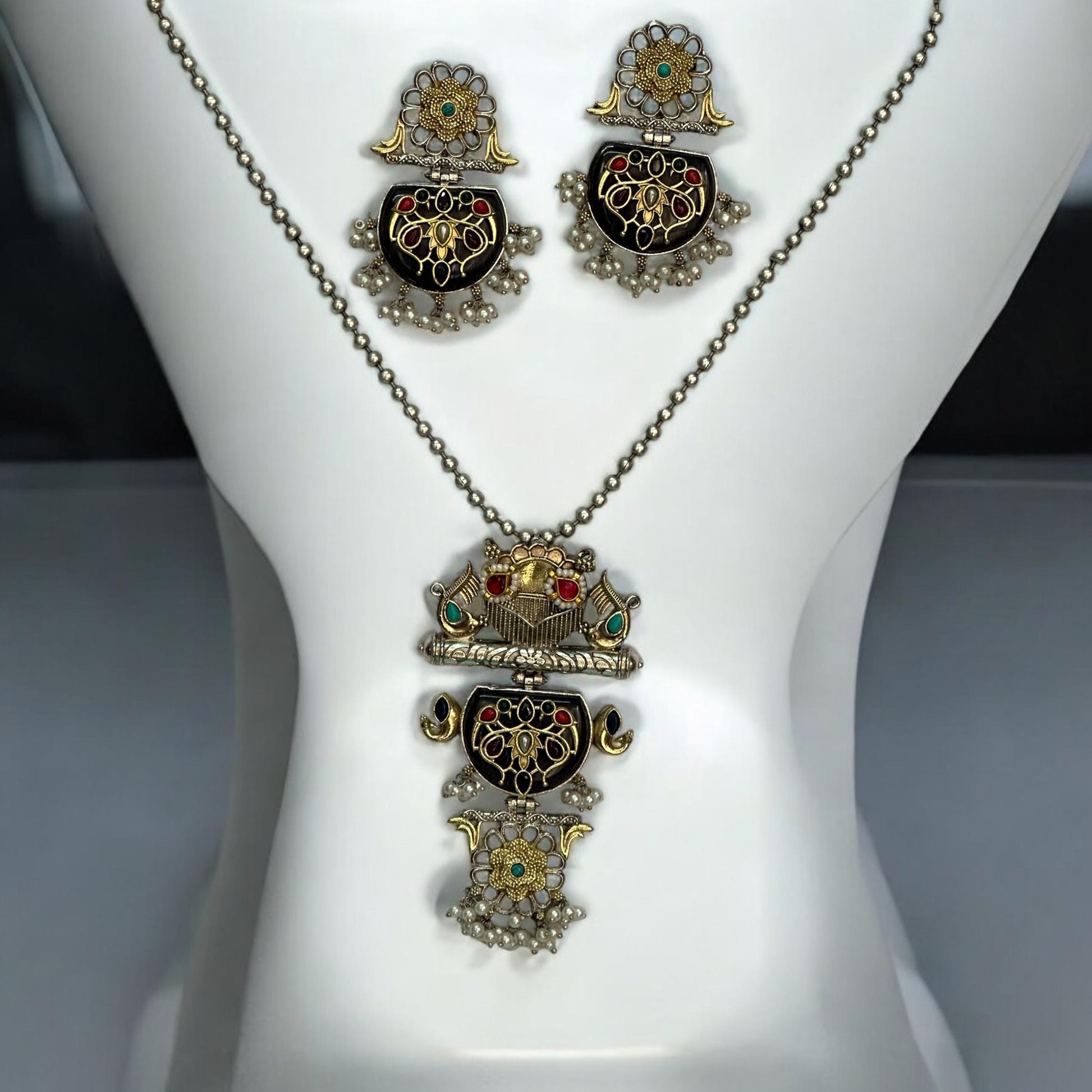 Dual Tone Apala Silver Long Pendant Set with Intricate Carving and Black Stone Finish, Featuring Matching Earrings placed on a white coloured jewellery display stand.