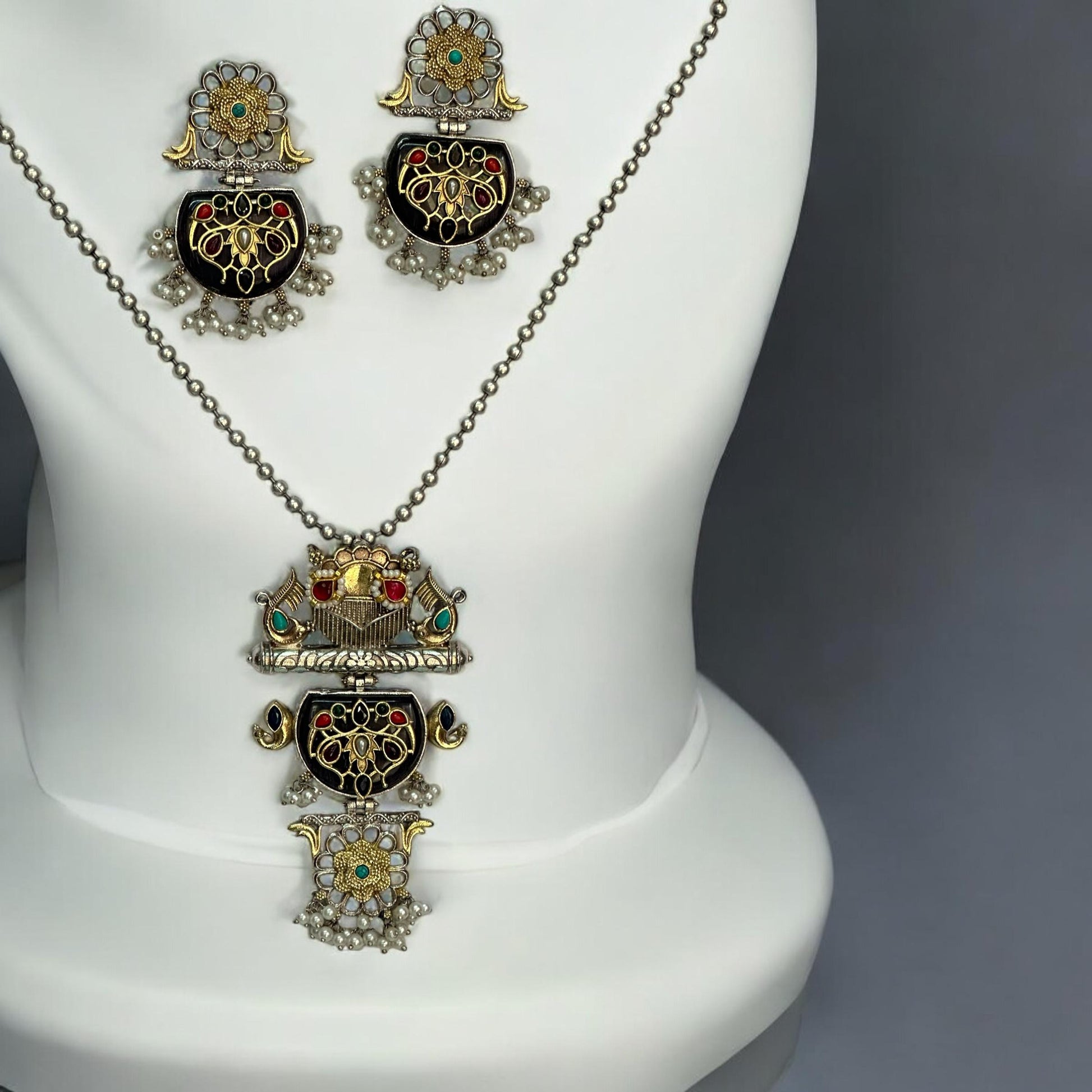 Dual Tone Apala Silver Long Pendant Set with Intricate Carving and Black Stone Finish, Featuring Matching Earrings placed on a white coloured jewellery display stand.