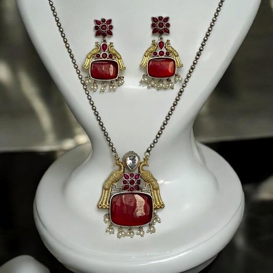 Dual Tone German Silver Long Chain Pendant Set with Red Stones and Matching Earrings, Featuring Intricate Traditional Design displayed on a white coloured jewellery display stand.
