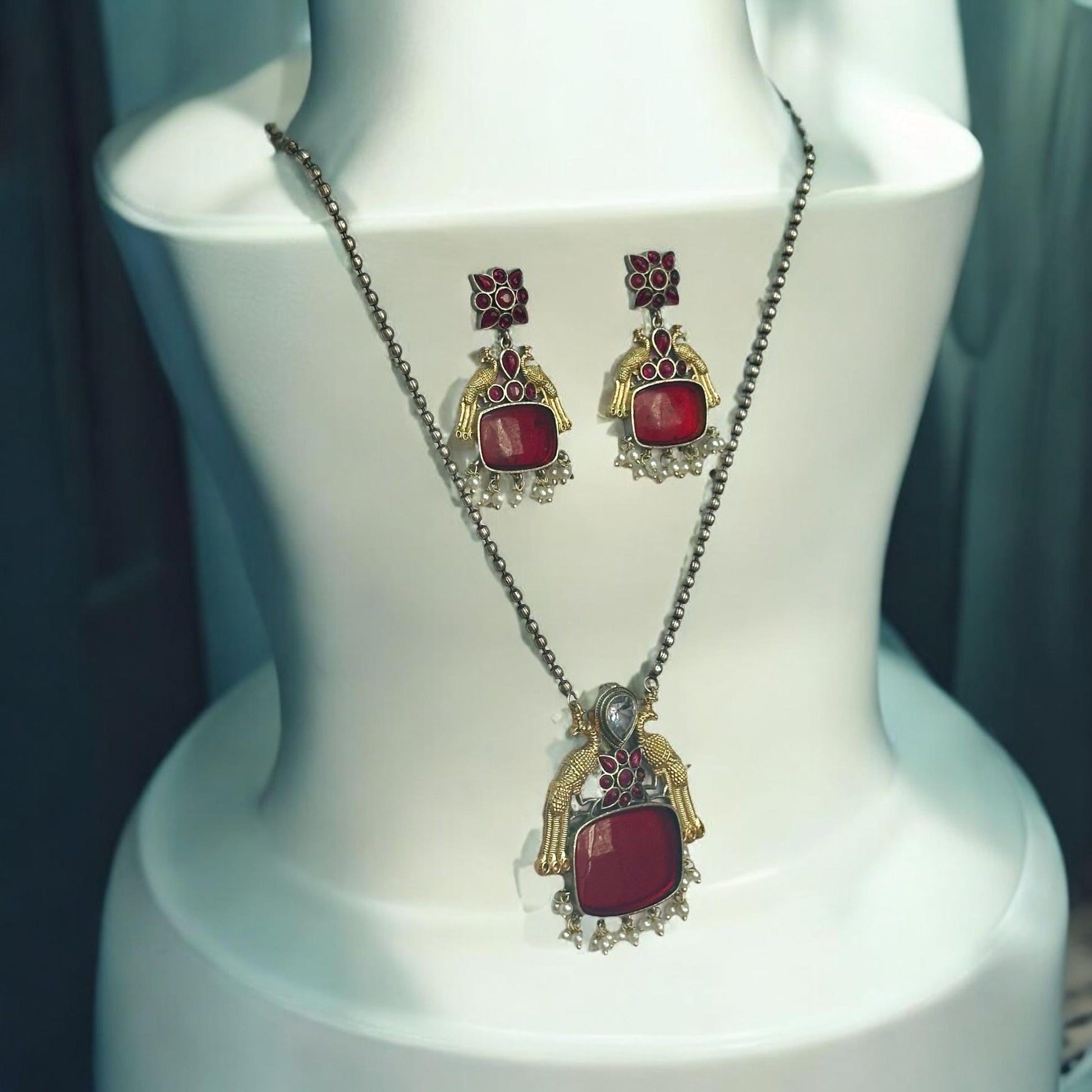 Dual Tone German Silver Long Chain Pendant Set with Red Stones and Matching Earrings, Featuring Intricate Traditional Design showcased on a white coloured jewellery display stand.
