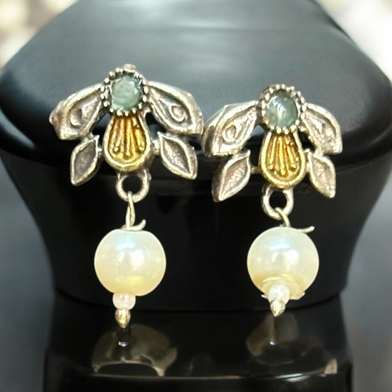 Dual-tone silver-look-alike earrings with intricate yellow and white detailing, accented with green enamel, displayed on a black mannequin with a soft light background.