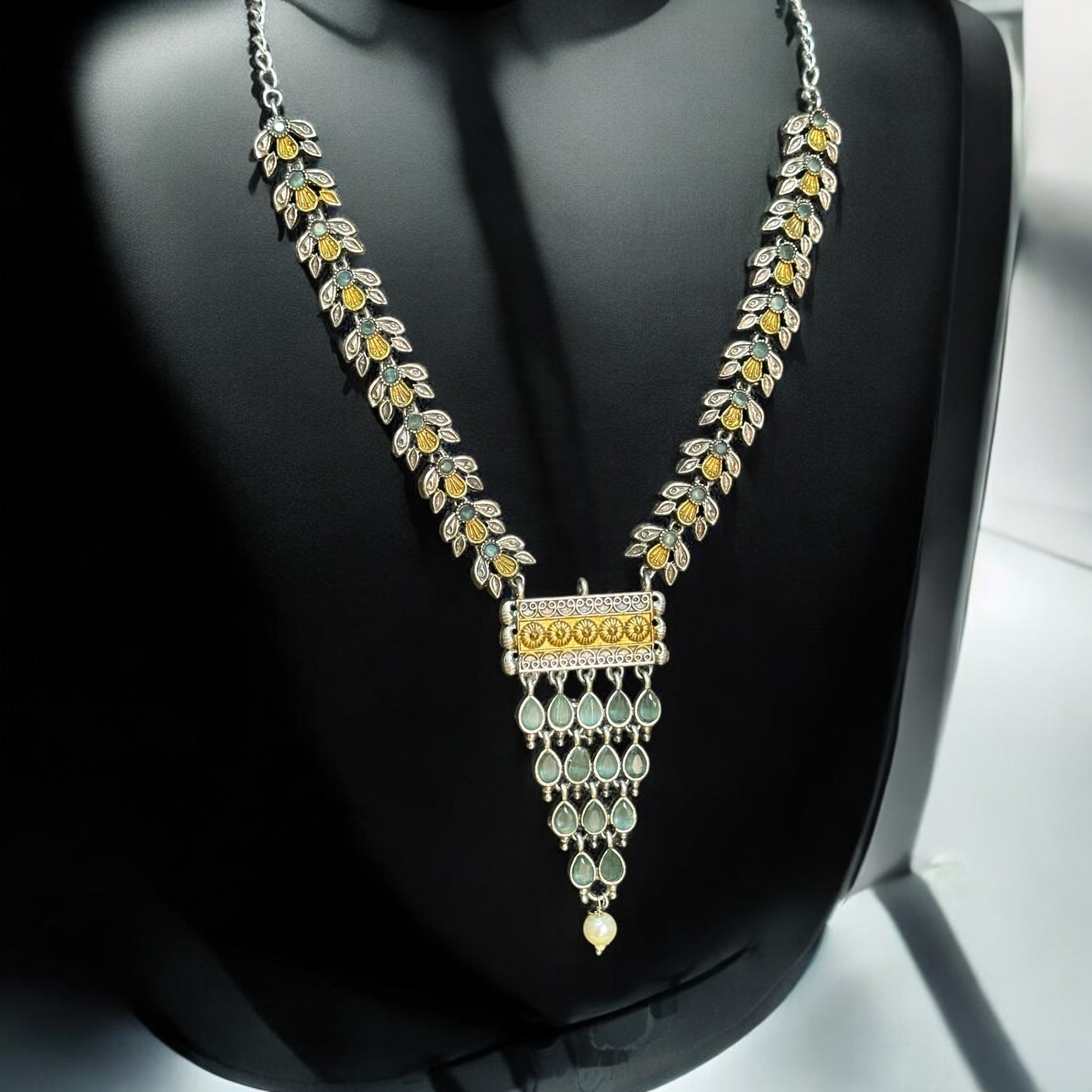 Dual-tone silver-look-alike necklace with intricate yellow and white detailing, accented with green enamel displayed on a black mannequin with a soft light background.