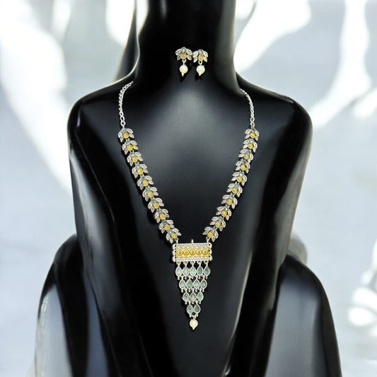 Dual-tone silver-look-alike necklace with intricate yellow and white detailing, accented with green enamel and matching earrings, displayed on a black mannequin with a soft light background.