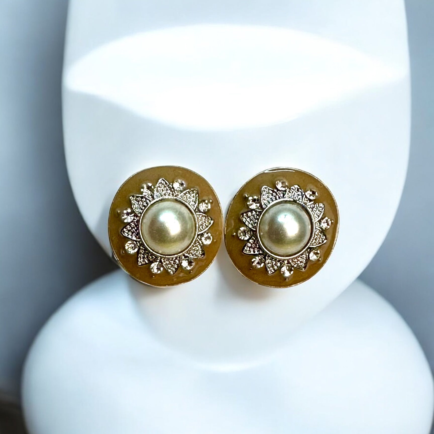 Elegant pearl stud earrings with a gold-toned circular base, featuring intricate floral detailing and embedded white stones, displayed on a white jewellery display stand with a soft blue backdrop.