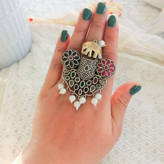 A Fusion Oxidised Adjustable Ring with intricate oxidised silver detailing, colorful gemstones, pearl drops, and an elephant motif, worn on a hand with green-painted nails against a lace fabric background.