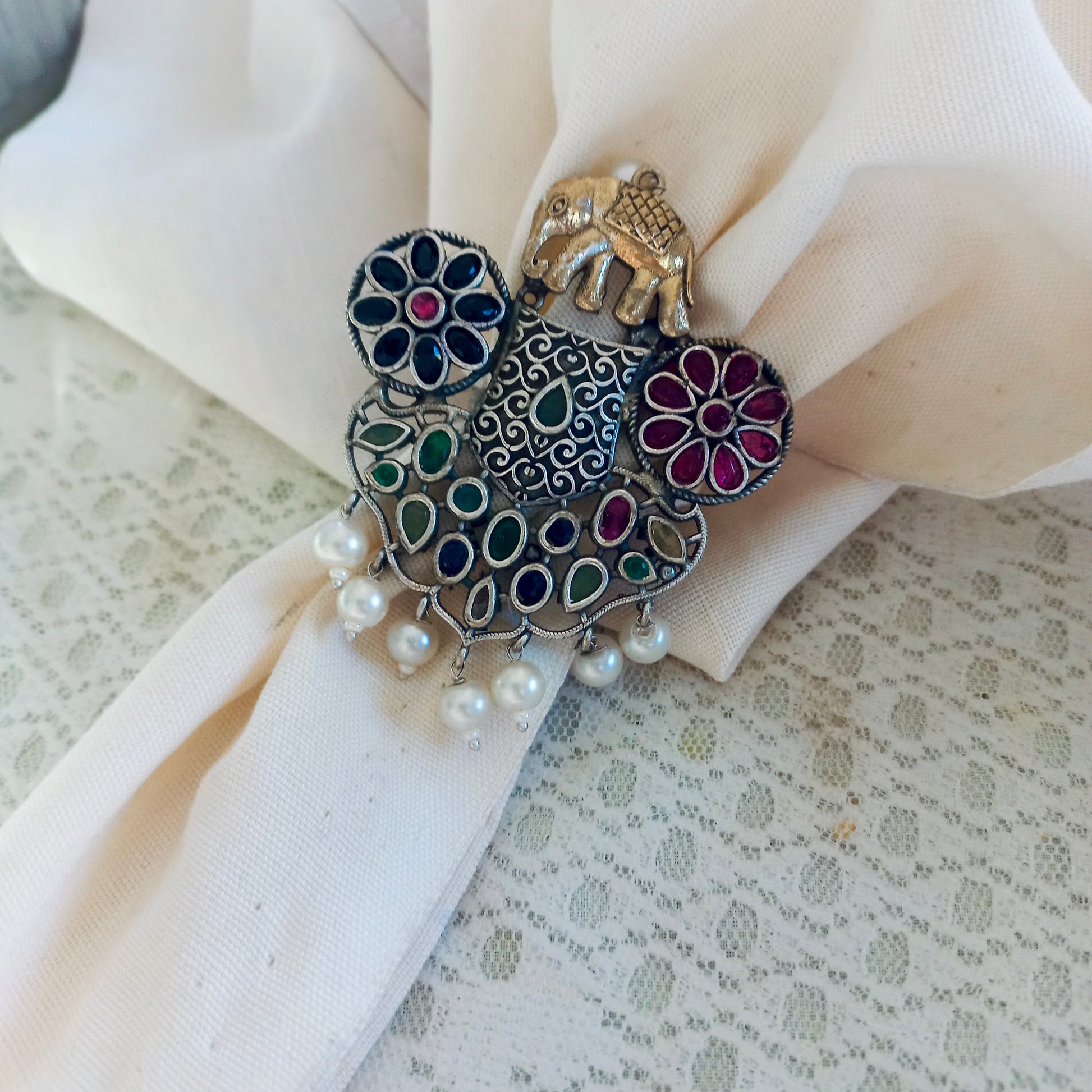 A Fusion Oxidised Adjustable Ring with intricate oxidised silver detailing, colorful gemstones, pearl drops, and an elephant motif,  against a lace fabric background.