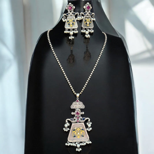 German silver replica necklace with an intricate pendant design featuring floral motifs in gold and pink highlights, paired with matching statement earrings, displayed on a black mannequin with a soft, elegant background.