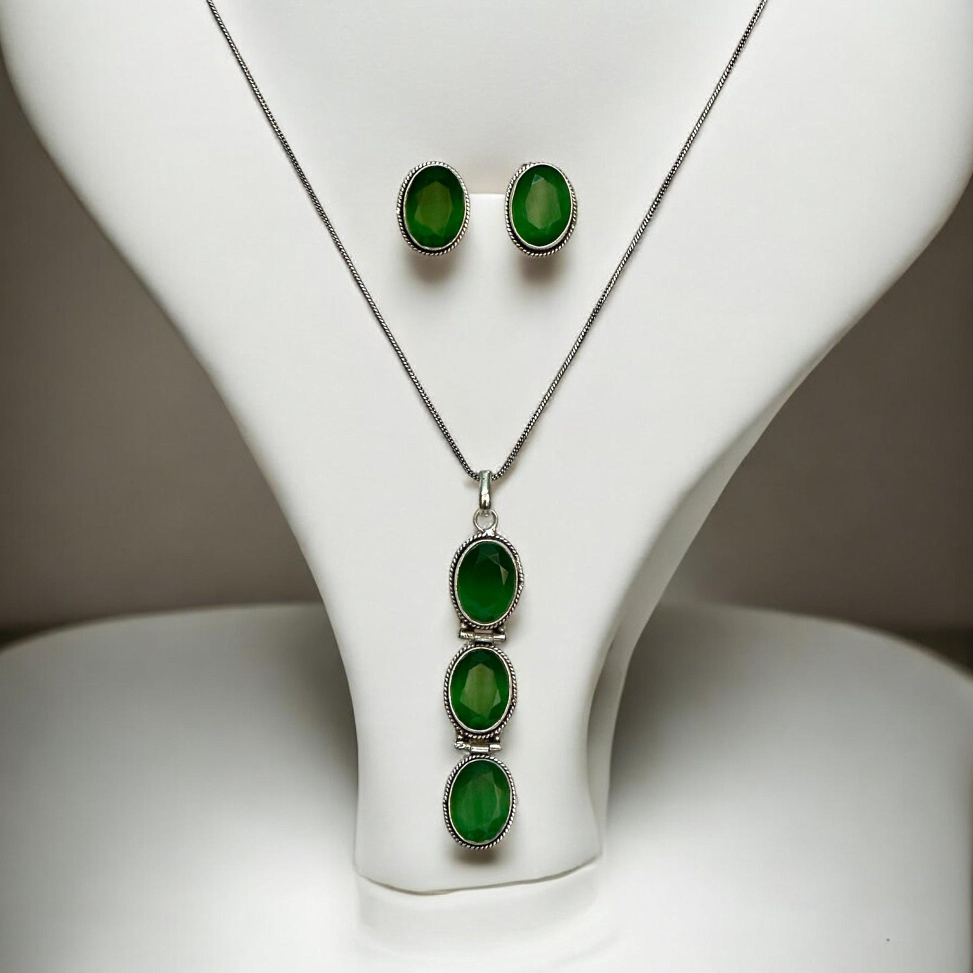 German Silver Long Chain Pendant Set featuring three vertically aligned green oval gemstones in a silver frame, paired with matching green stud earrings, displayed on a white jewellery display stand with minimalist background.