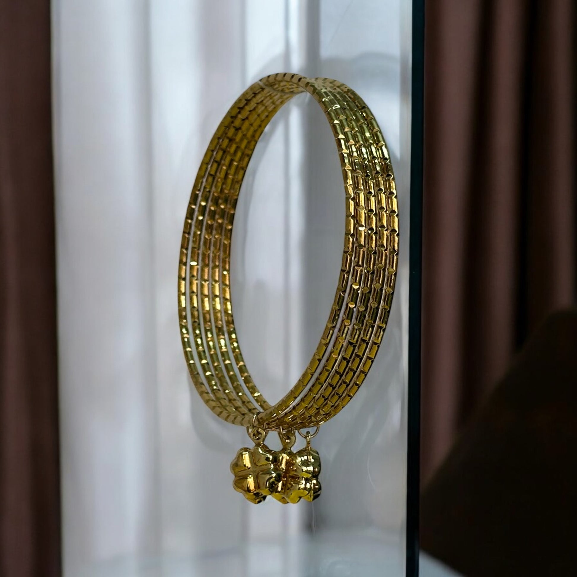 Golden bangle set featuring delicate ghungroo charms, ideal for traditional and festive occasions. Hanging with a background of white and brown curtains.