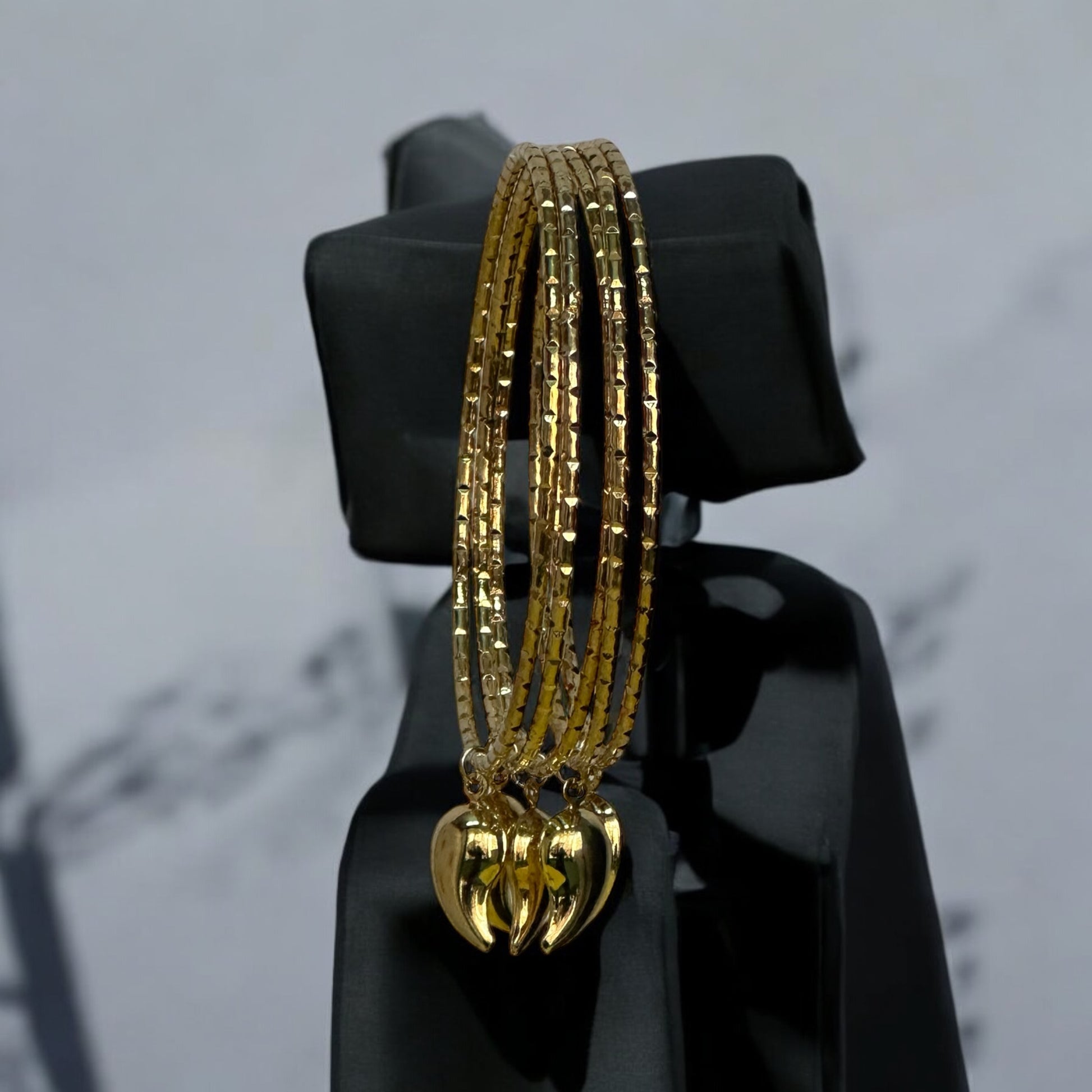 Golden bangle set featuring intricate patterns and adorned with hanging charms, displayed on a black jewellery display stand with a soft gray background.