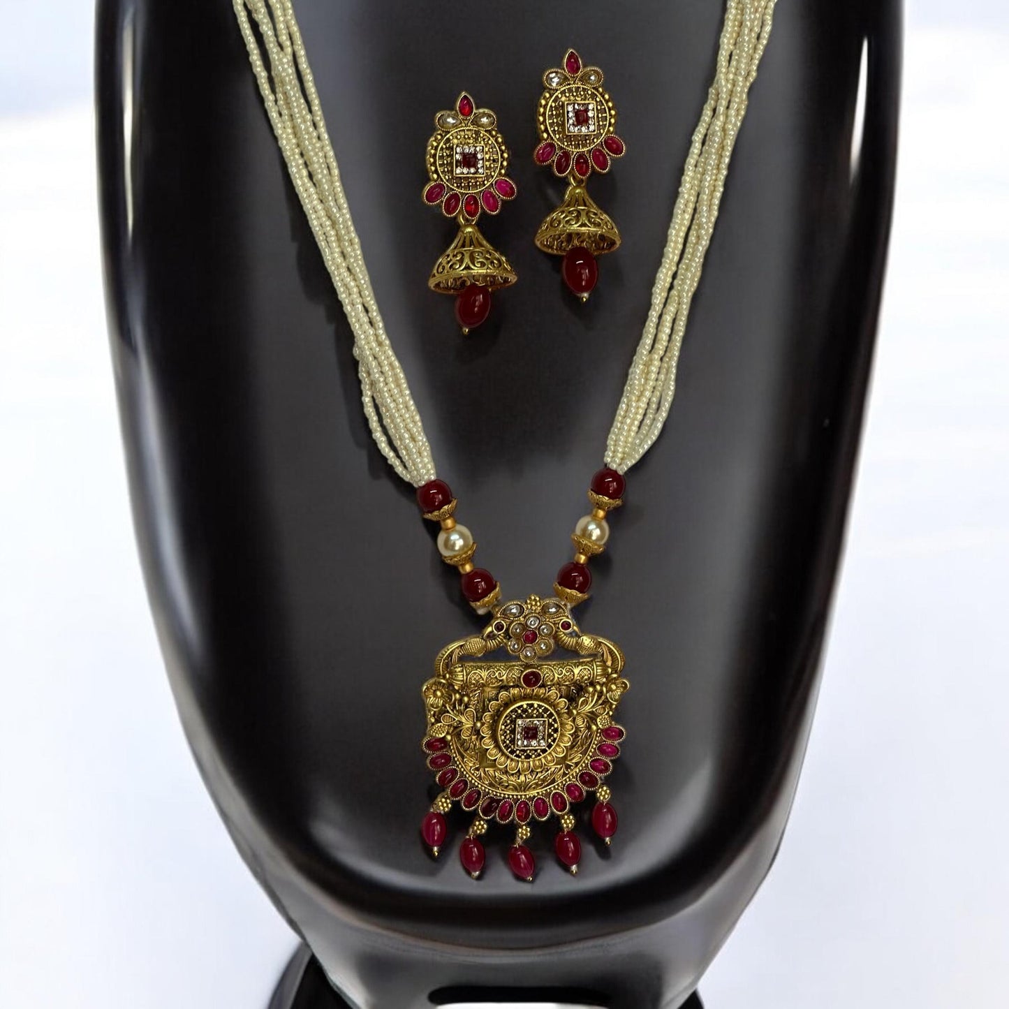 Traditional gold-toned long necklace set featuring a detailed pendant with red stones and pearl bead chains, paired with matching dangler earrings, displayed on a black mannequin with white background.