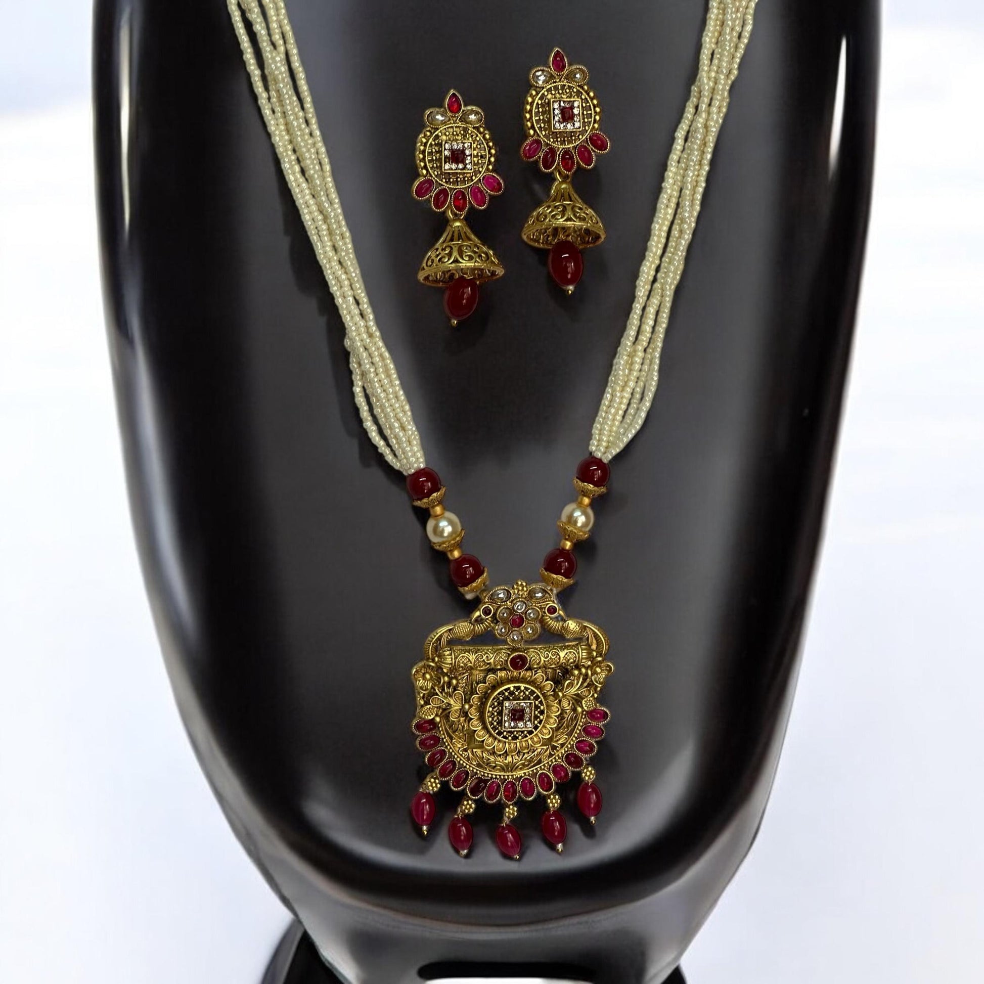 Traditional gold-toned long necklace set featuring a detailed pendant with red stones and pearl bead chains, paired with matching dangler earrings, displayed on a black mannequin with white background.