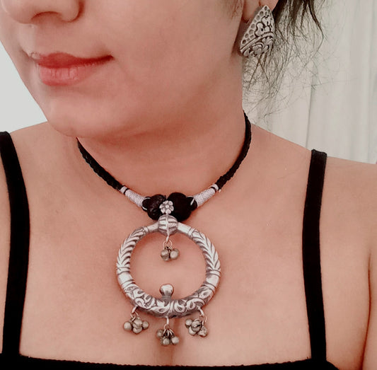 A model displaying Handmade silver replica necklace featuring a bold circular pendant with intricate engravings and ghungroo charms, paired with detailed triangular stud earrings. 