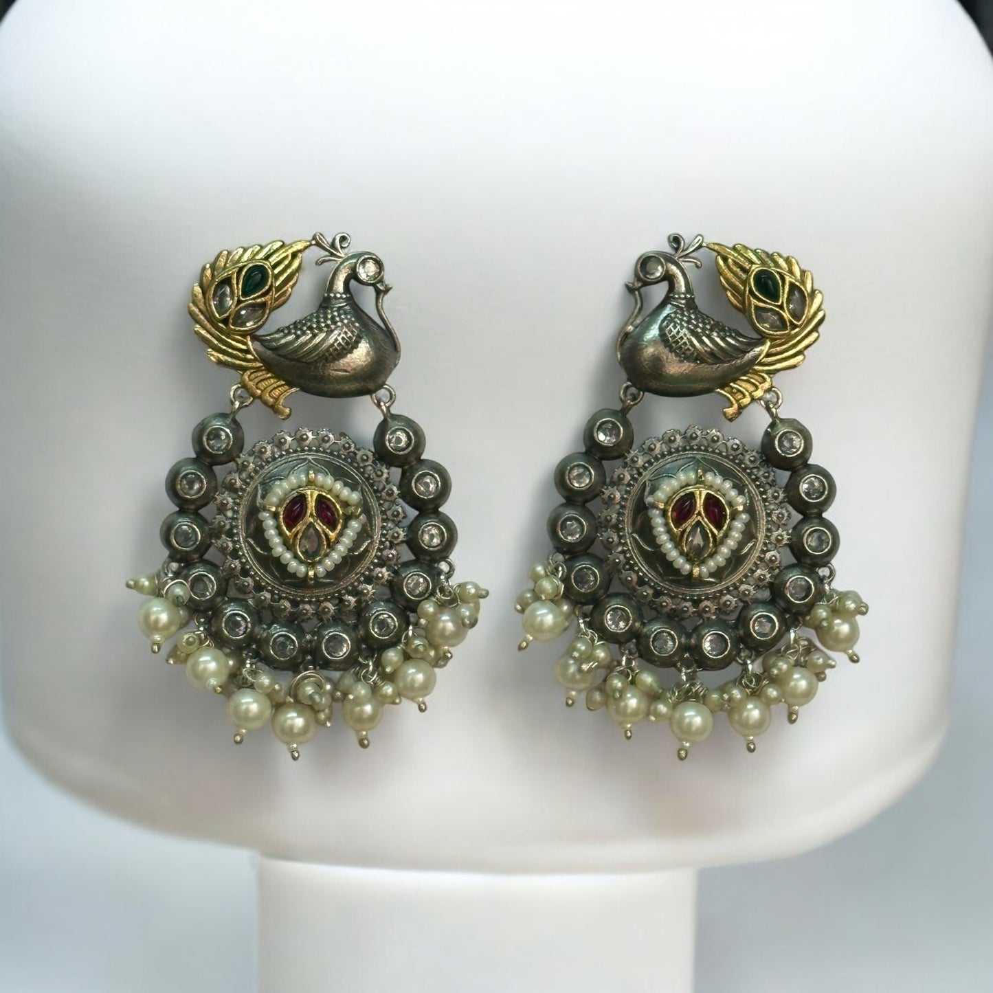 Jadau Kundan dual-tone statement earrings featuring peacock motifs, intricate circular detailing, red and green enamel work, and pearl hangings, displayed on an off white jewelry stand with a soft blue background.