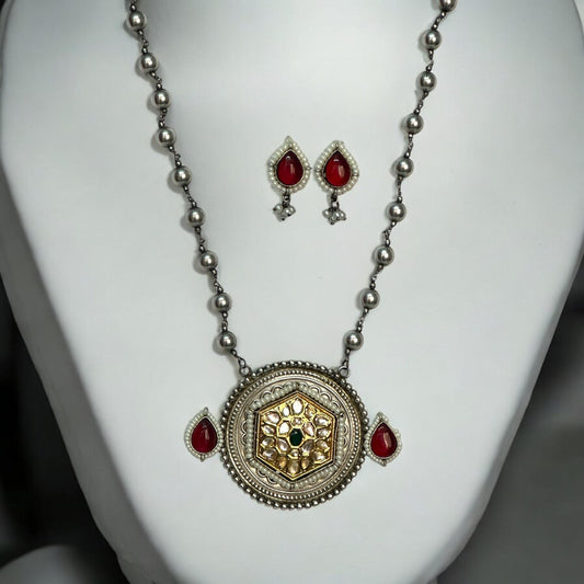 Dual tone Apala-style big-sized pendant set featuring a gold and silver finish with Kundan work, paired with red and green gemstone accents, displayed on a white mannequin.